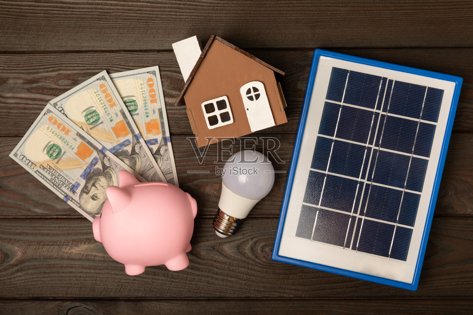 在棕色的木头上，用太阳能电池板、led灯、房屋模型、钱和存钱罐组成的平面构图。节约资金和清洁能源的概念。生态与可持续发展的理念。照片摄影图片