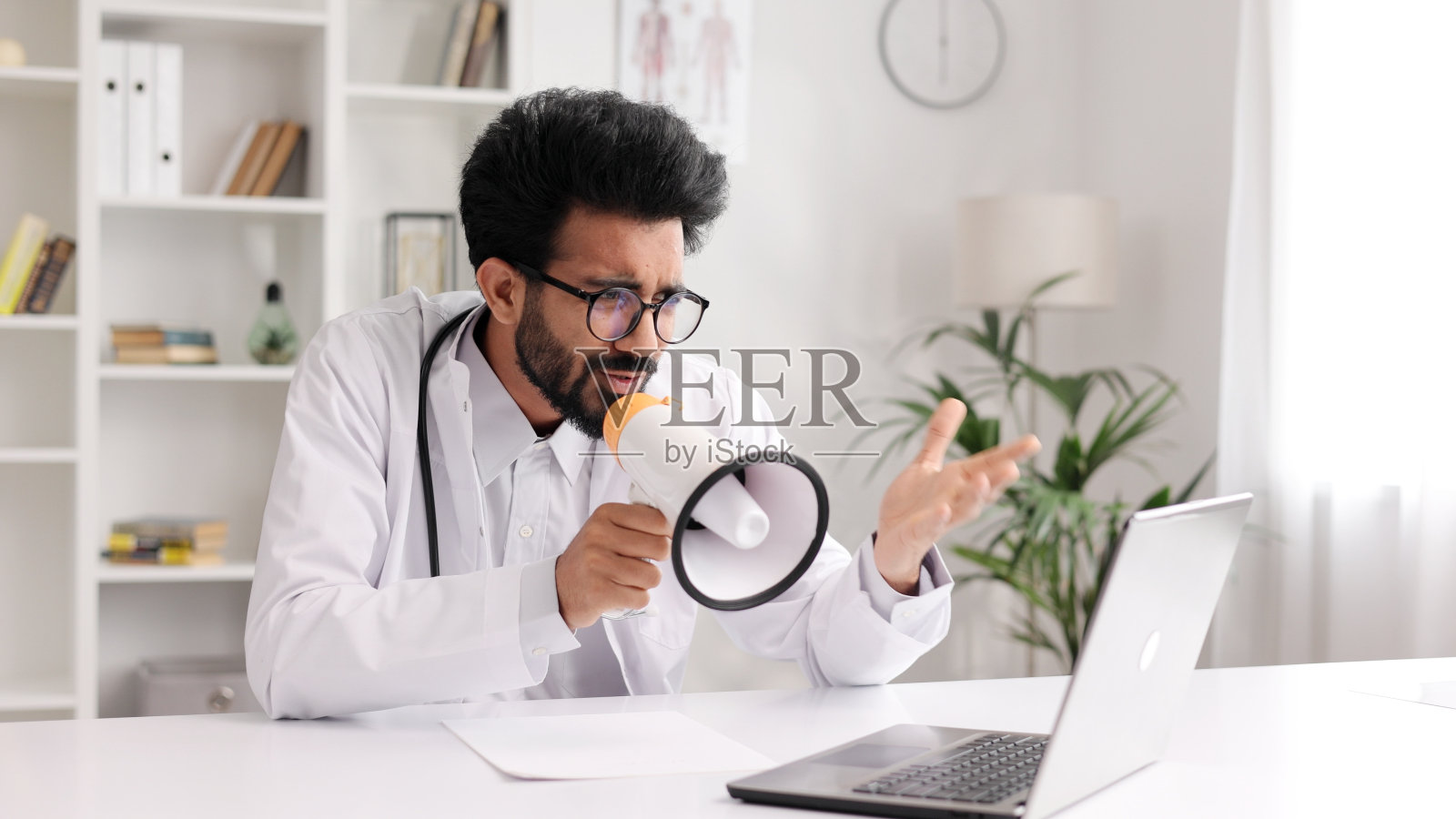 一位穿着白大褂的印度男医生试图向不听他说话的病人传达他的意见，他对着扬声器大喊。夸大情绪，警告危险的疾病。照片摄影图片