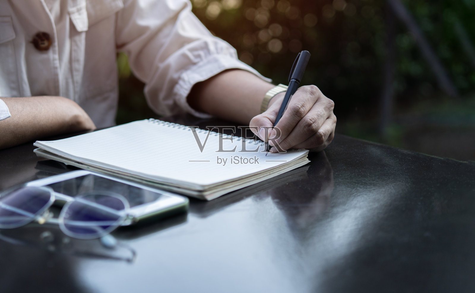 Crop anonymous male writing notes with pen in notebook while sitting in the garden near in soft light.照片摄影图片