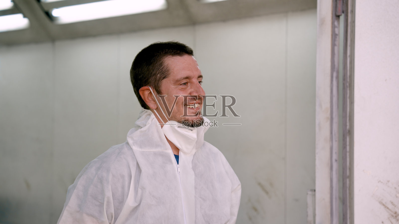 穿着白色防护服的白人男性站在五个工厂标准汽车喷漆车间。戴着防雾面罩和护目镜，我听到他在修车时双臂交叉。照片摄影图片