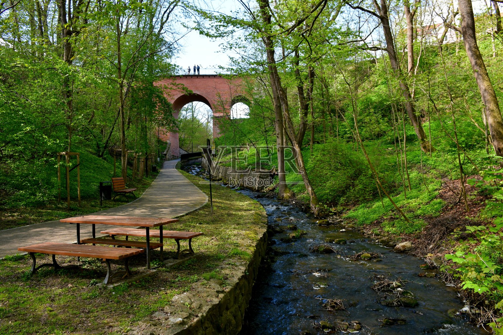 一个小湖泊或小溪流经一个山谷，山谷里长满了树木、草药、鲜花和其他种类的植物，远处可以看到一座红砖桥，前面有一张桌子照片摄影图片