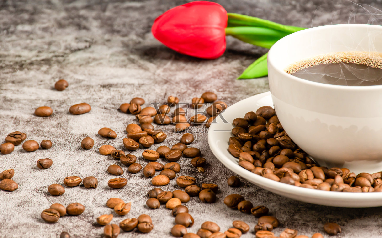 水泥地板上的桌子上放着一个白色杯子里的热咖啡特写，咖啡杯碟里放着许多烤过的咖啡豆，周围，烟雾和香气从杯子里飘出来。模糊的背景照片摄影图片