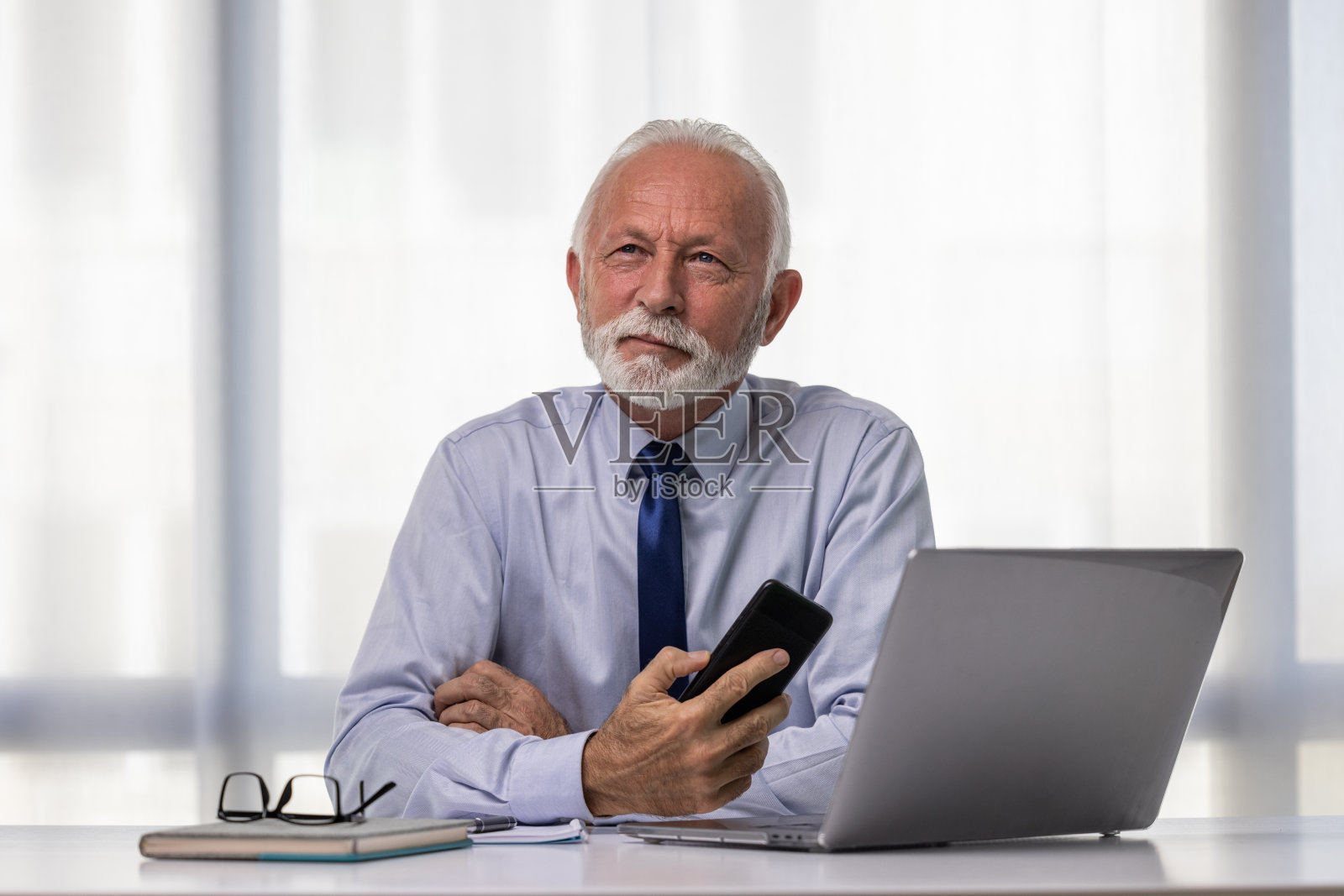 快乐的资深商人在他现代化的办公室里工作。成熟的CEO或董事坐在桌子旁，用笔记本电脑和手机，看着别处，面带微笑。照片摄影图片
