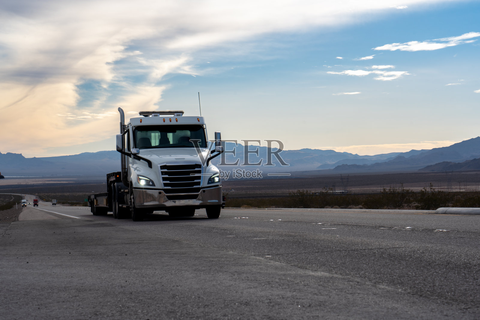 一辆白色的半挂车巡游在内华达沙漠，它巨大的车架与落日的金色形成鲜明的对比。高耸的群山构成了风景如画的背景。照片摄影图片