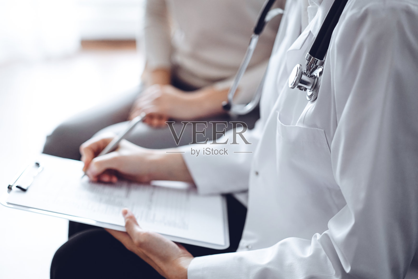 医生和病人坐在诊所办公室的沙发上。重点是女医生的手填写用药史记录表，近距离。医学的概念照片摄影图片