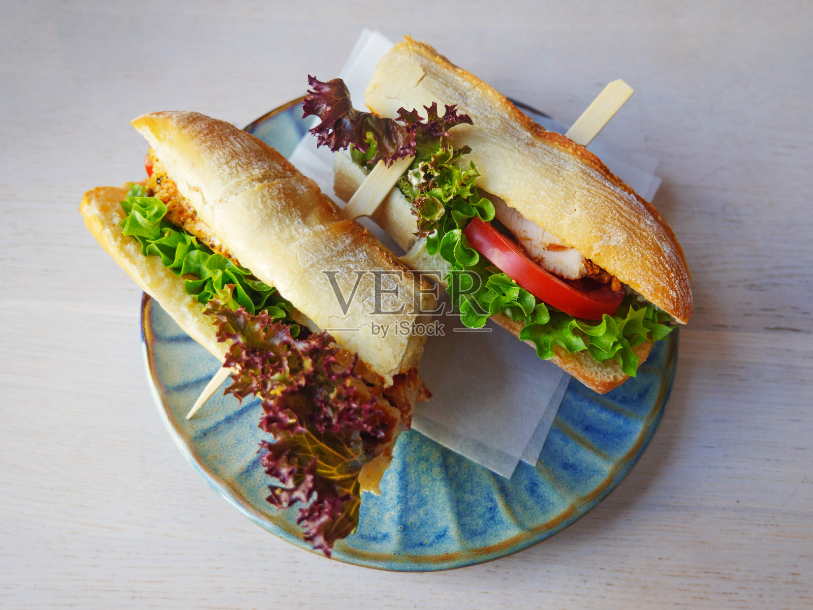 切片法棍三明治，夹着炸鸡、西红柿、生菜和酱汁，放在蓝色的彩色盘子上，咖啡馆餐厅的早餐俯视图照片摄影图片