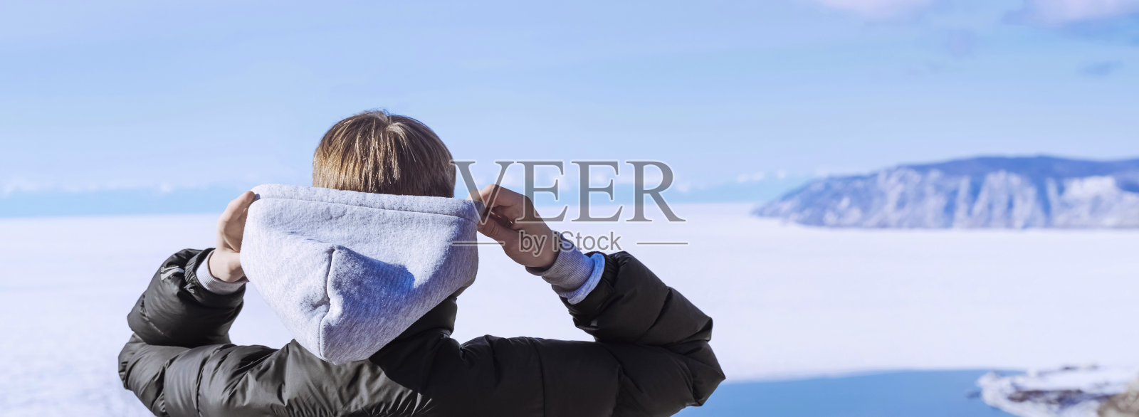 戴着兜帽的少年，穿着保暖衣服的青年旅行者站在那里欣赏大自然。在冬天欣赏白雪皑皑的海或湖的景色。照片摄影图片