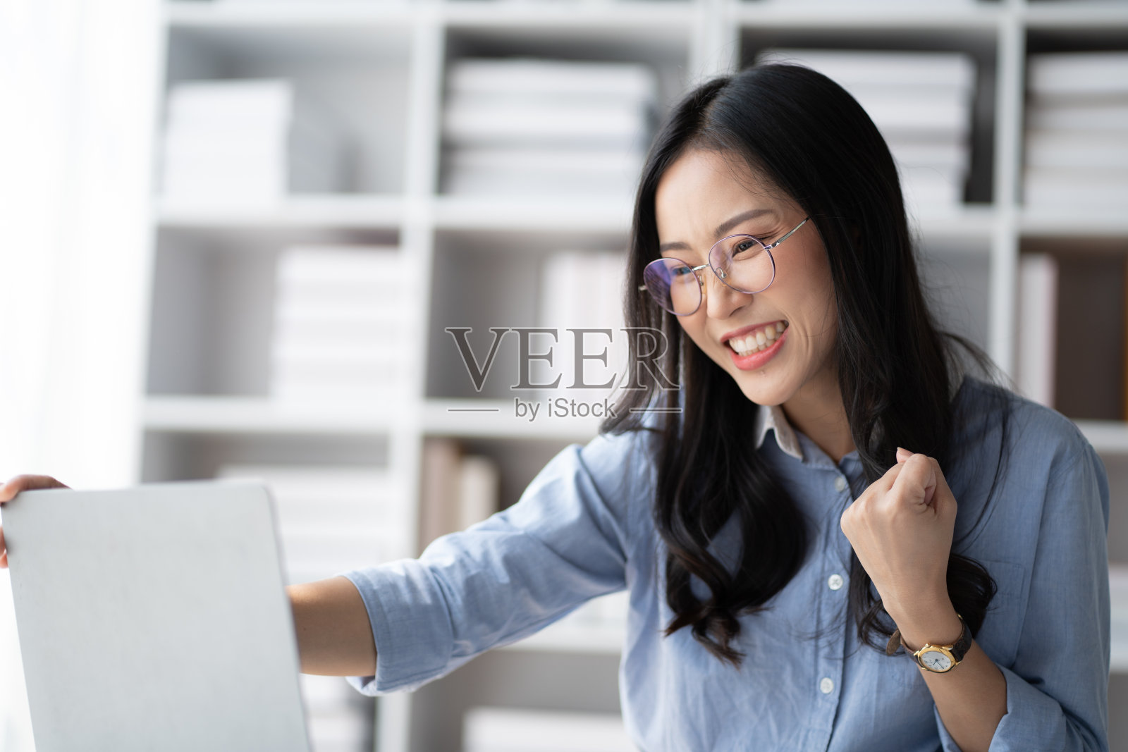 戴眼镜的亚洲女商人欣喜若狂，拿着笔记本电脑庆祝成功。照片摄影图片
