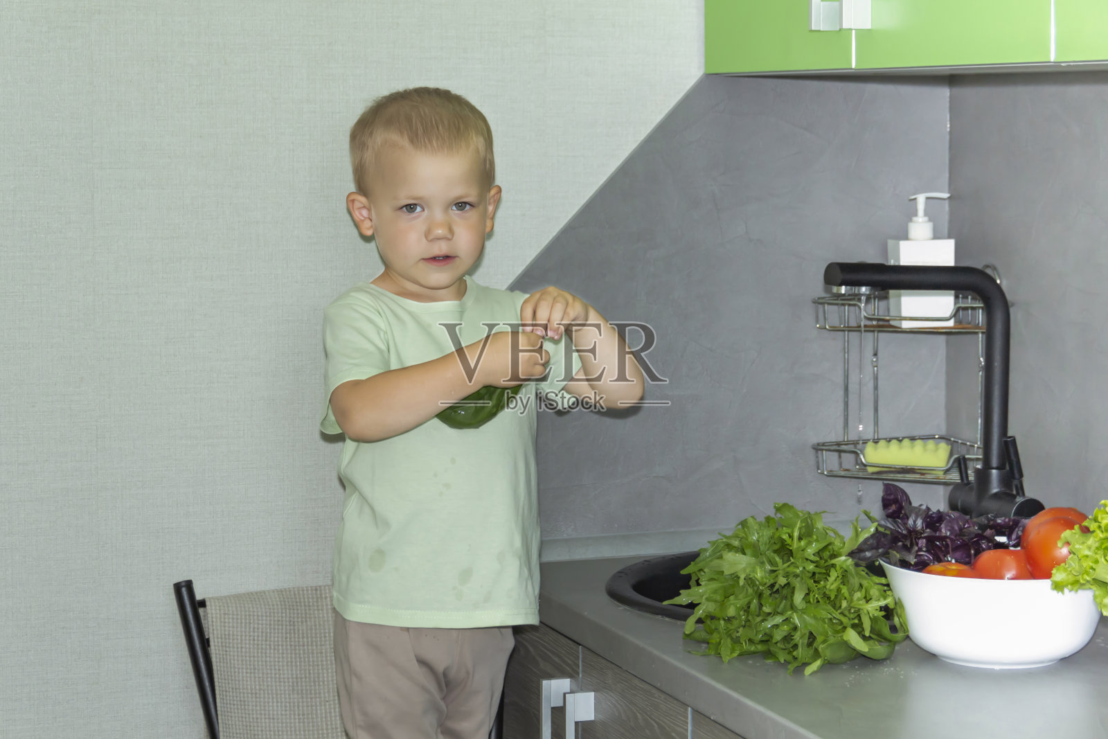 一个短发的小男孩在厨房帮忙做饭。在灰色厨房的黑色水槽里清洗新鲜的蔬菜和香草。照片摄影图片