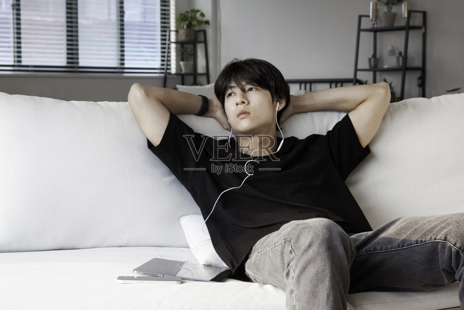 感觉孤独。沉思的年轻人坐在沙发上，手放在头上，看着窗外，用耳机听音乐。照片摄影图片