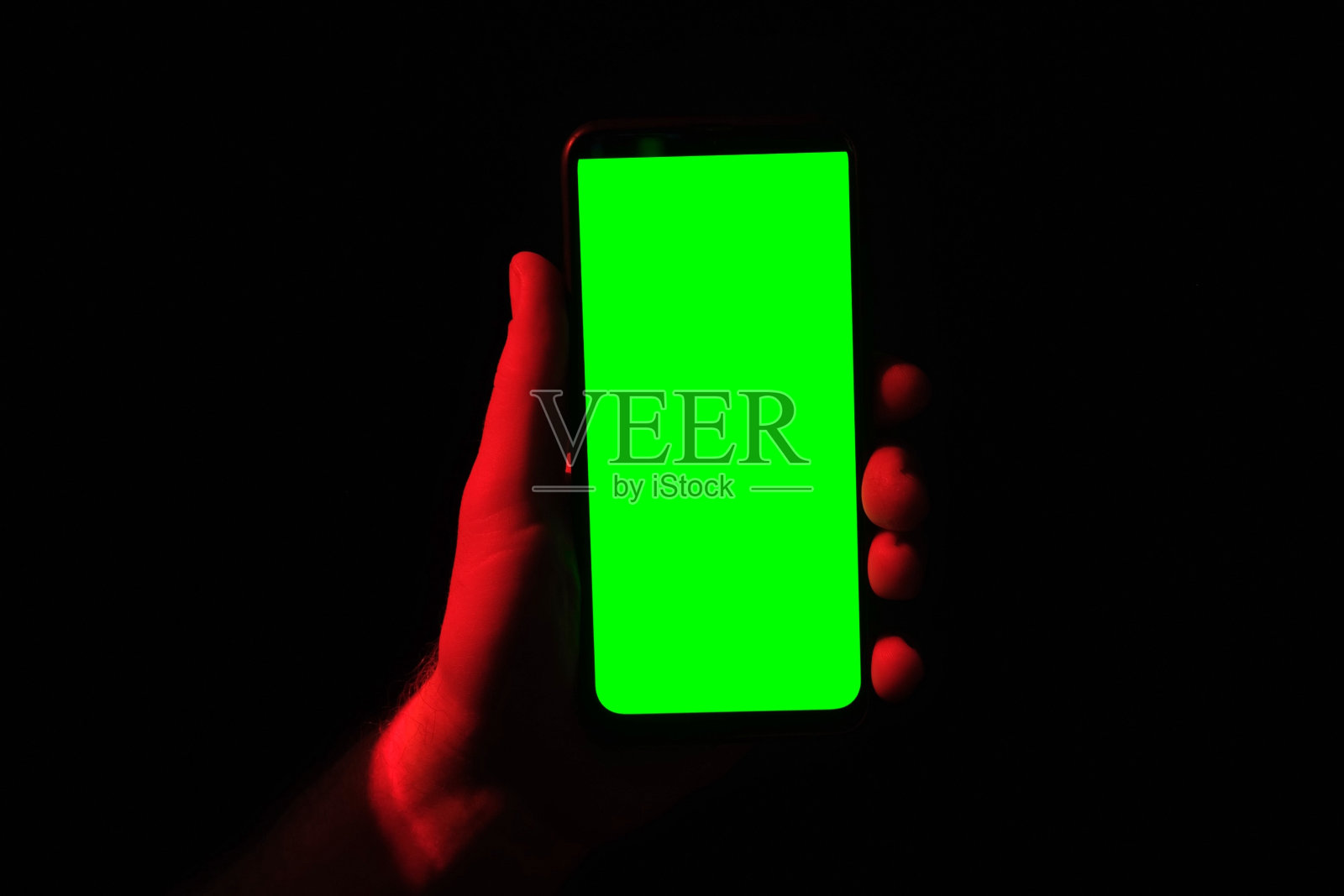 年轻人手里拿着手机。使用绿屏的智能手机配红蓝霓虹灯，背景暗。男性显示黑色手机色度键显示。关闭了。手抬起设备，保持它不动照片摄影图片