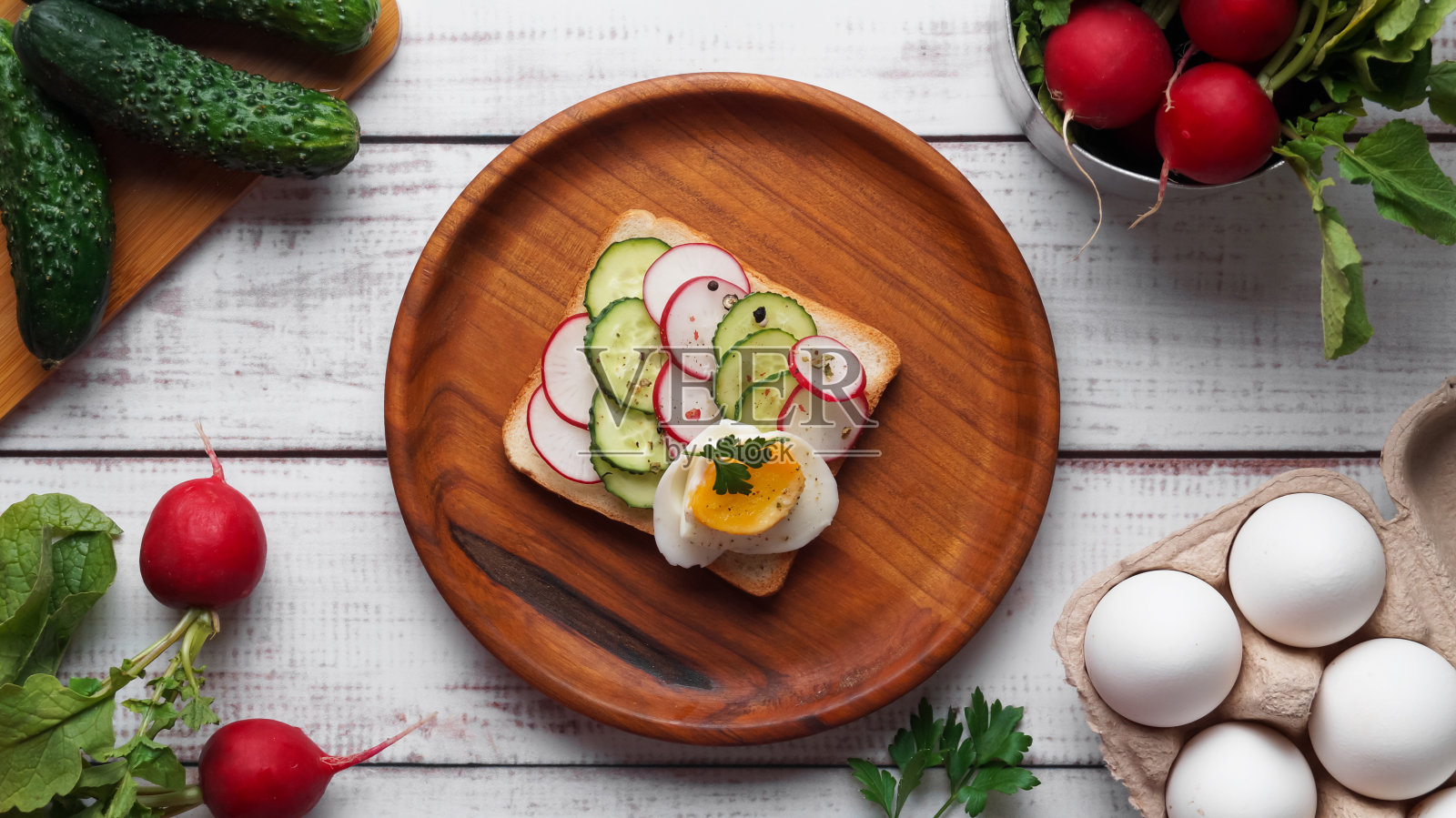 白色的木桌上平铺着煮鸡蛋、萝卜、黄瓜和配料的美味三明治照片摄影图片