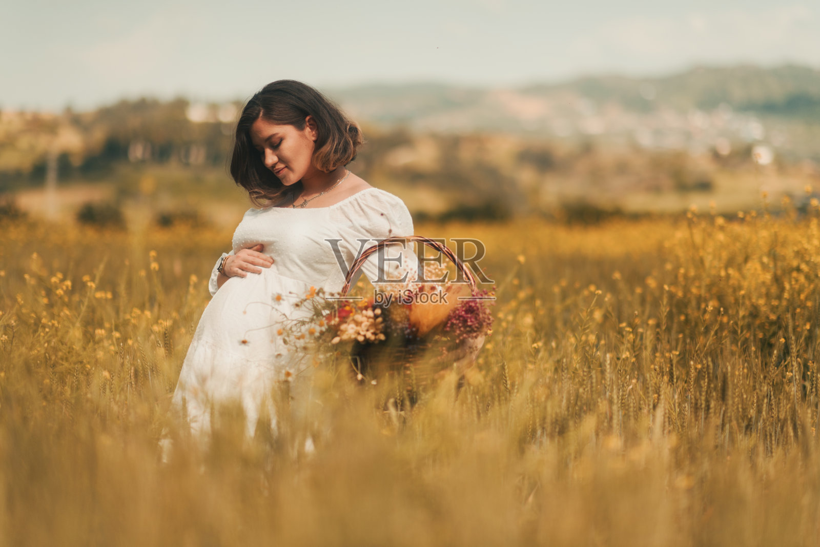 一位身穿白色长裙的孕妇提着一篮干花站在金色的麦田里，象征着母亲节的精神照片摄影图片