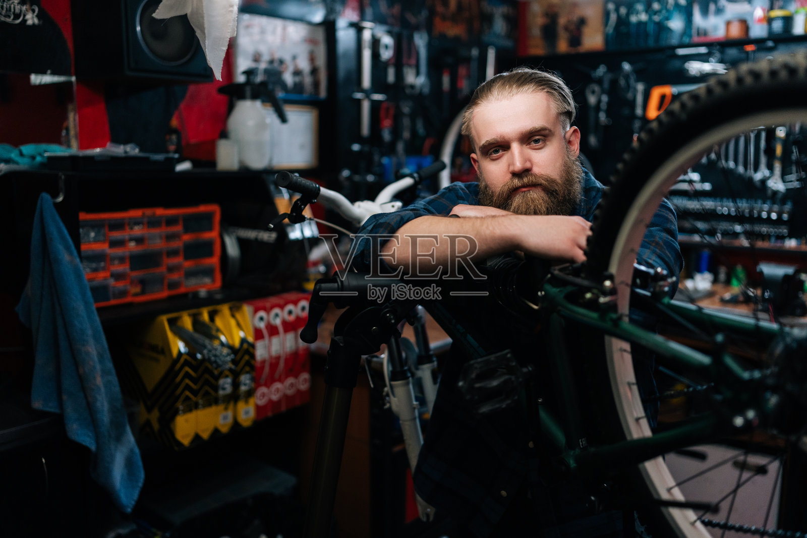 中镜头的肖像，英俊的自行车修理工有胡子站在自行车上靠在修理自行车车间黑暗的内部，看着相机。照片摄影图片