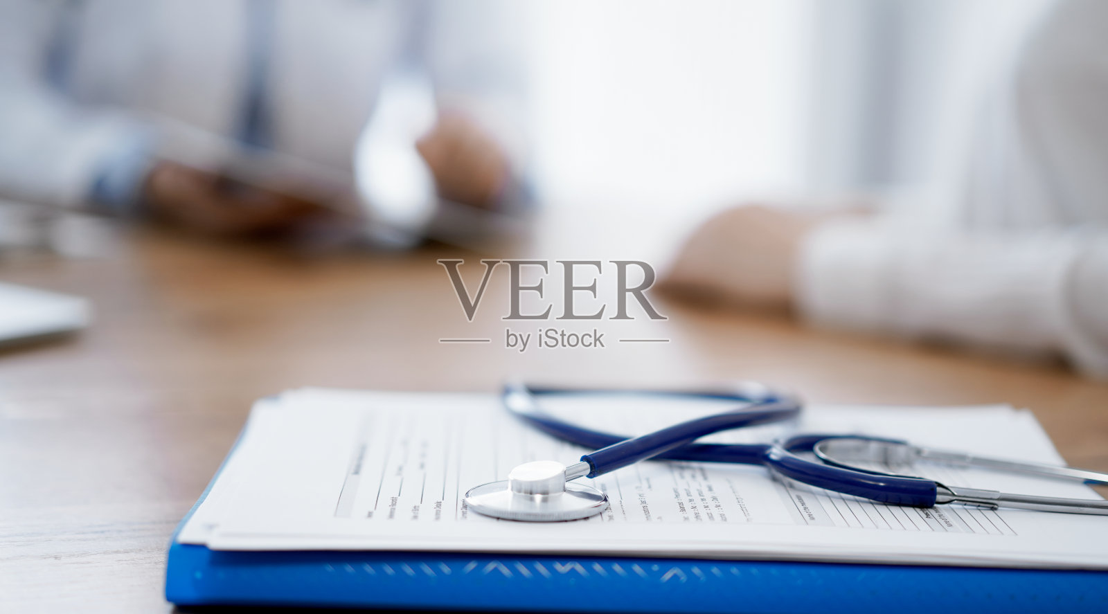 木桌上放着听诊器和用药史记录表，背景是医患双方坐着交谈。医学照片摄影图片