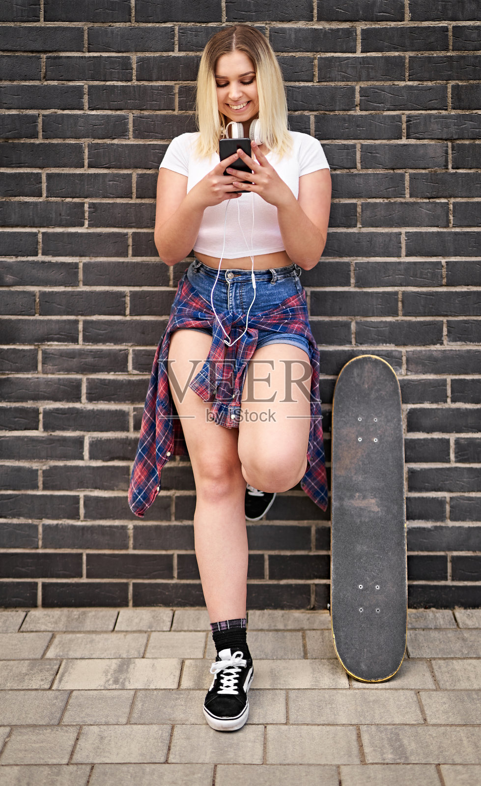 快乐的年轻开朗的千禧滑板学生女孩站在一堵黑砖墙前，用她的智能手机聊天-人们的生活方式和科技概念与青少年照片摄影图片