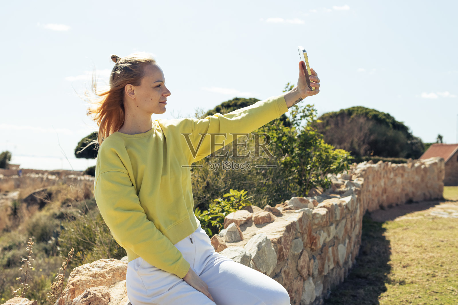 一个美丽的女孩坐在有石头的篱笆上，对着蓝天自拍。美丽的风景和一个女孩照片摄影图片