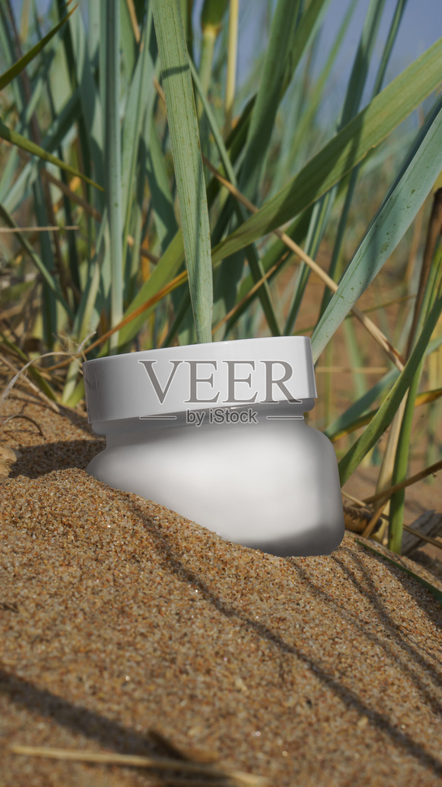 奶油罐模型。一个白色磨砂玻璃罐躺在沙滩上，周围是明亮的阳光下的植物照片摄影图片