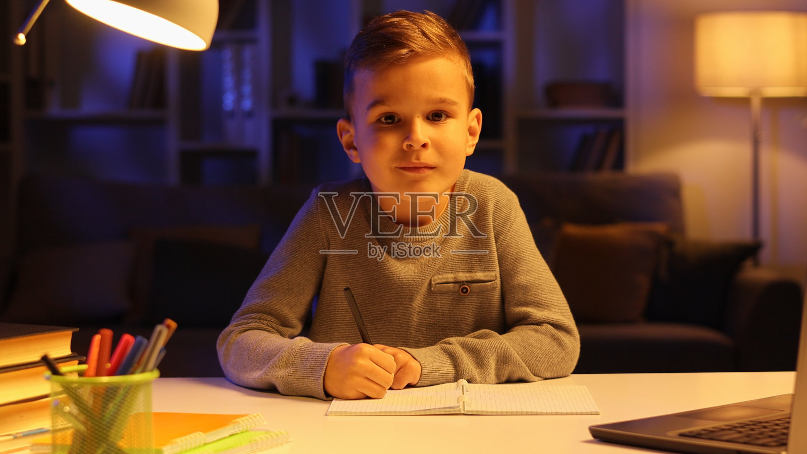 一个晚上做作业的小男孩的肖像，看着镜头，微笑着。照片摄影图片