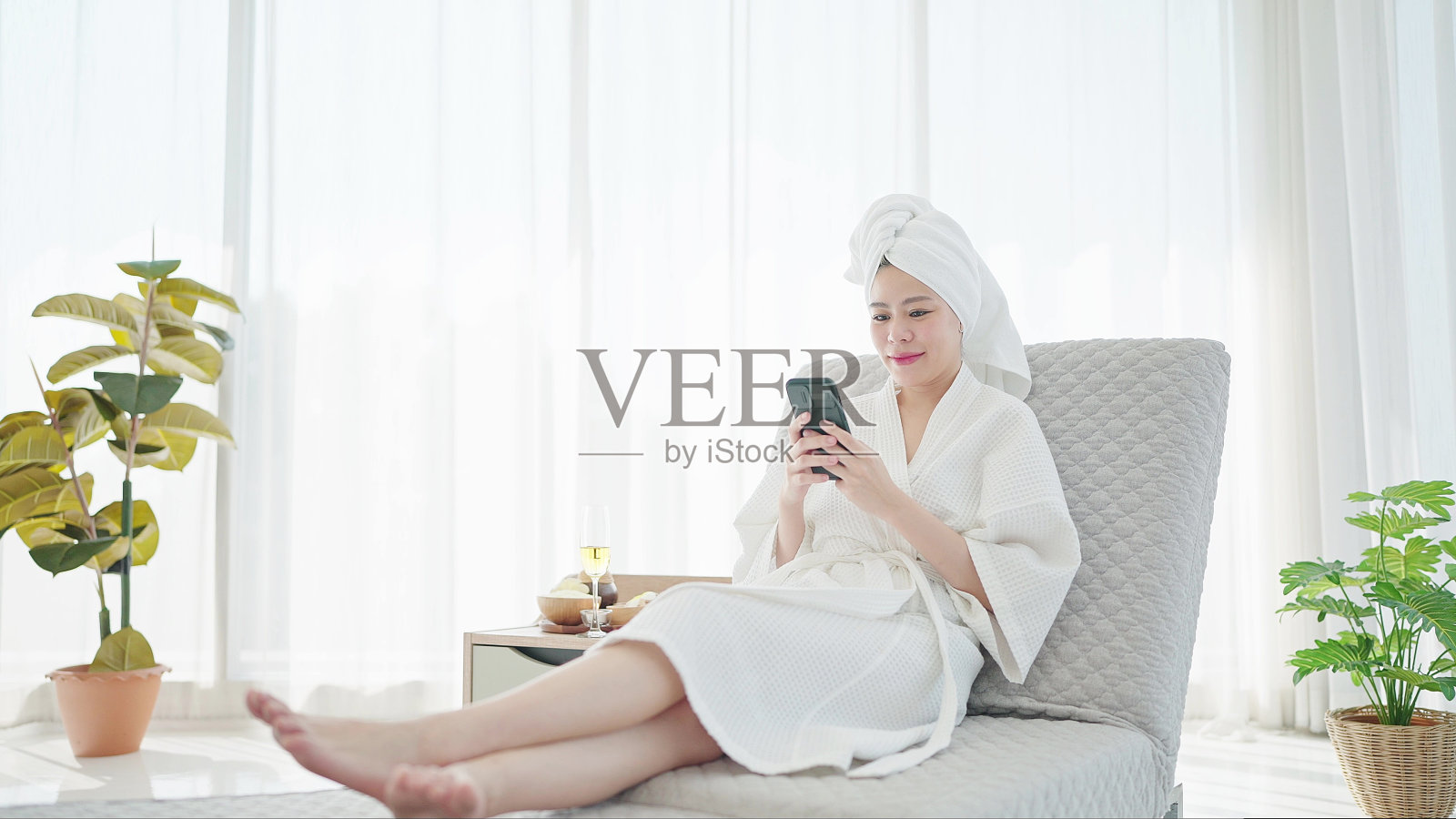 漂亮的年轻亚洲女人穿着浴袍和毛巾头巾，在现代客厅的沙发床上放松地使用智能手机照片摄影图片