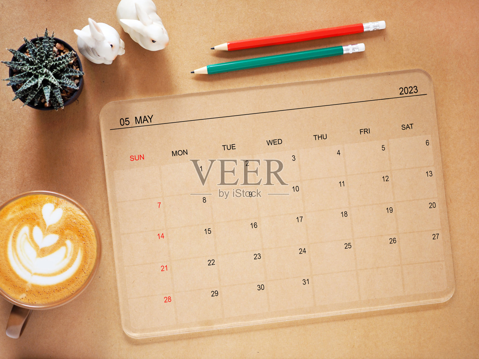 俯视图，透明的平板电脑显示2023年5月的日历应用程序页面和仙人掌，咖啡杯，铅笔，兔子雕像在棕色背景。照片摄影图片