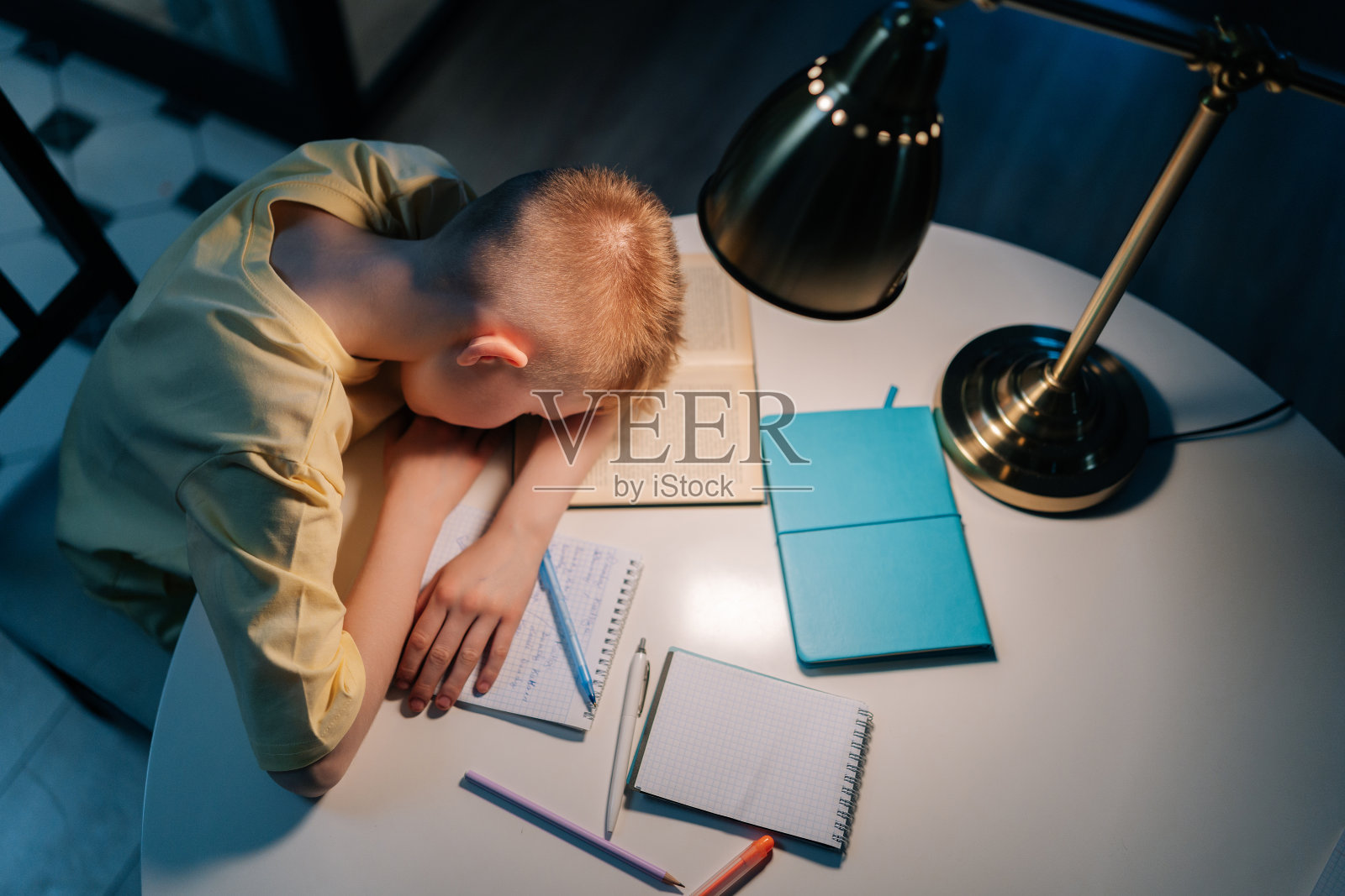 俯视图:在昏暗的房间里，在灯光下，一个疲惫的小男孩带着书、练习本和课本睡在桌子旁。照片摄影图片