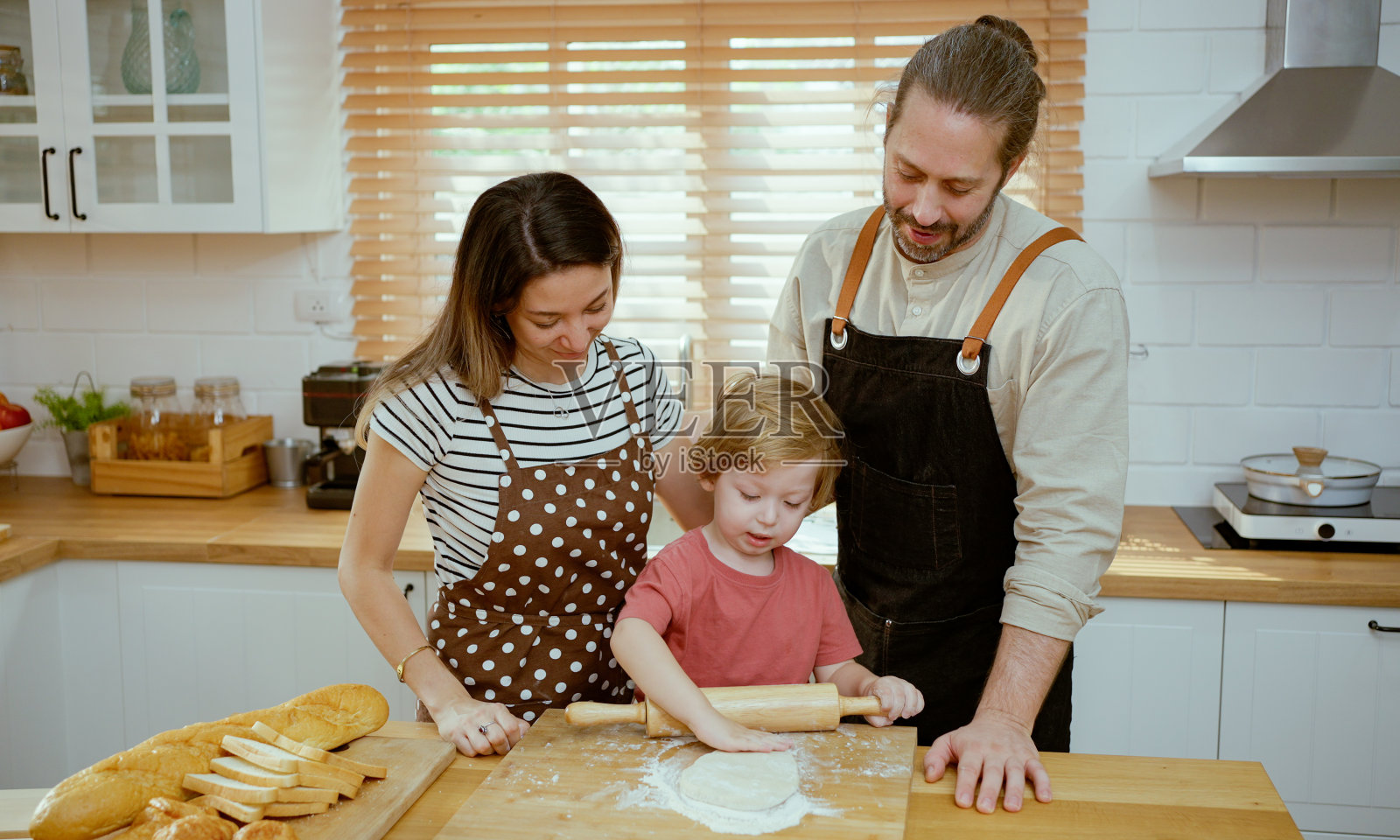 父亲和母亲在家里的厨房柜台上教小儿子揉面。父母和孩子一起享受室内烹饪的乐趣。照片摄影图片