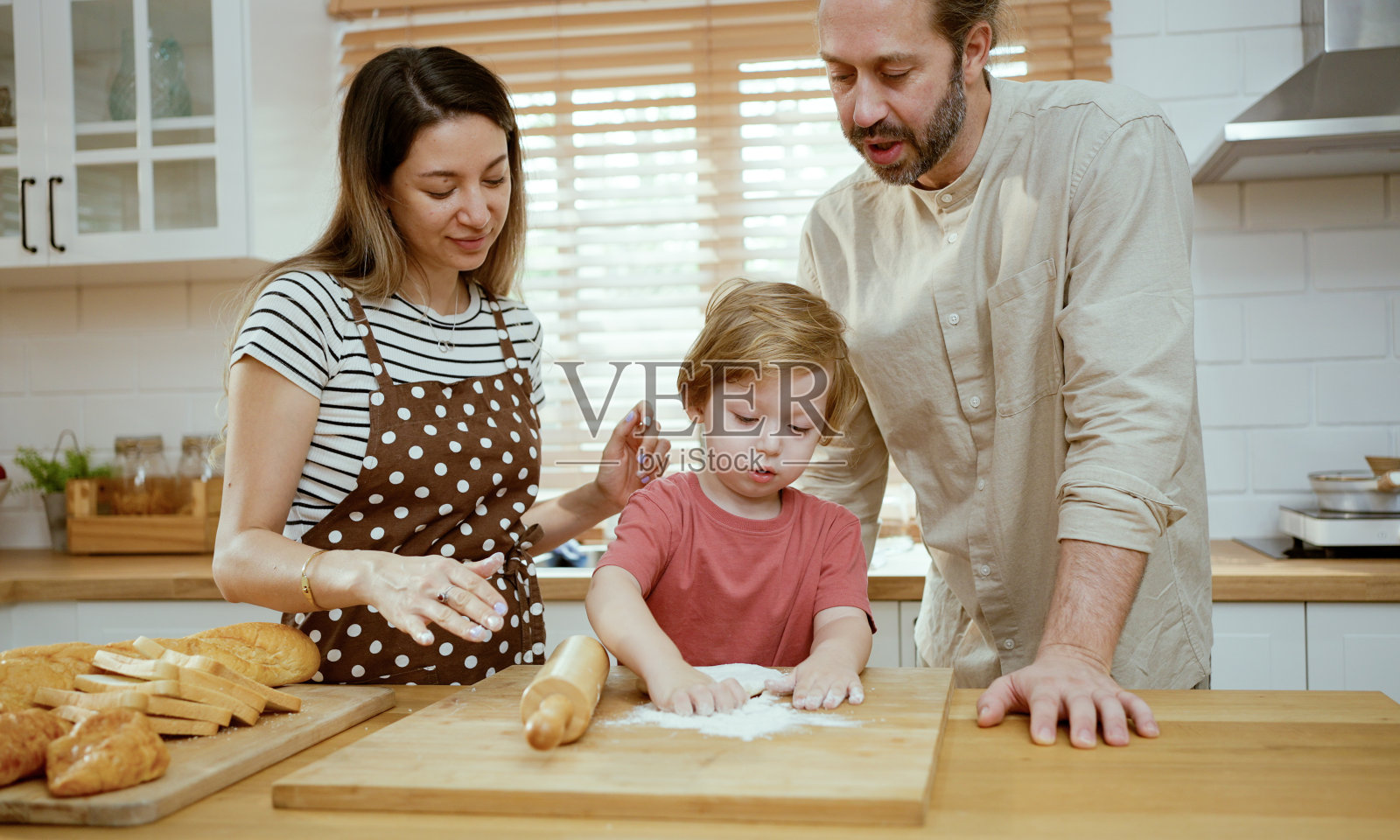 父亲和母亲在家里的厨房柜台上教小儿子揉面。父母和孩子一起享受室内烹饪的乐趣。照片摄影图片