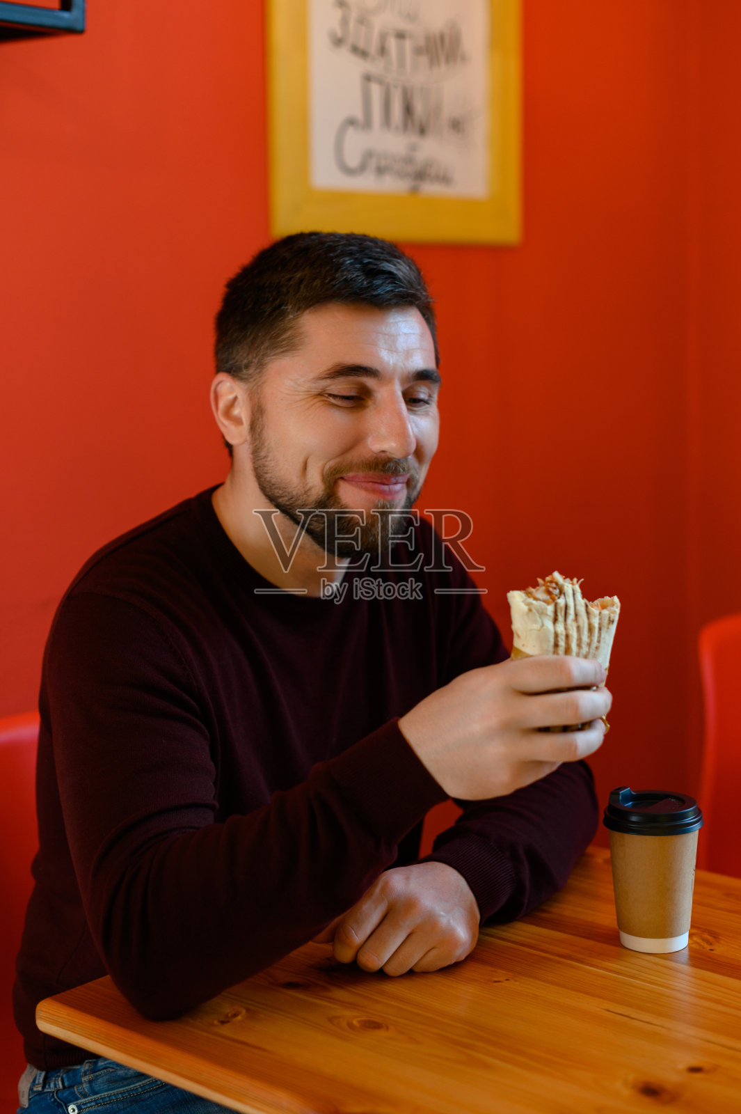 一个男人坐在咖啡馆里，面带微笑，手里拿着沙瓦玛。照片摄影图片
