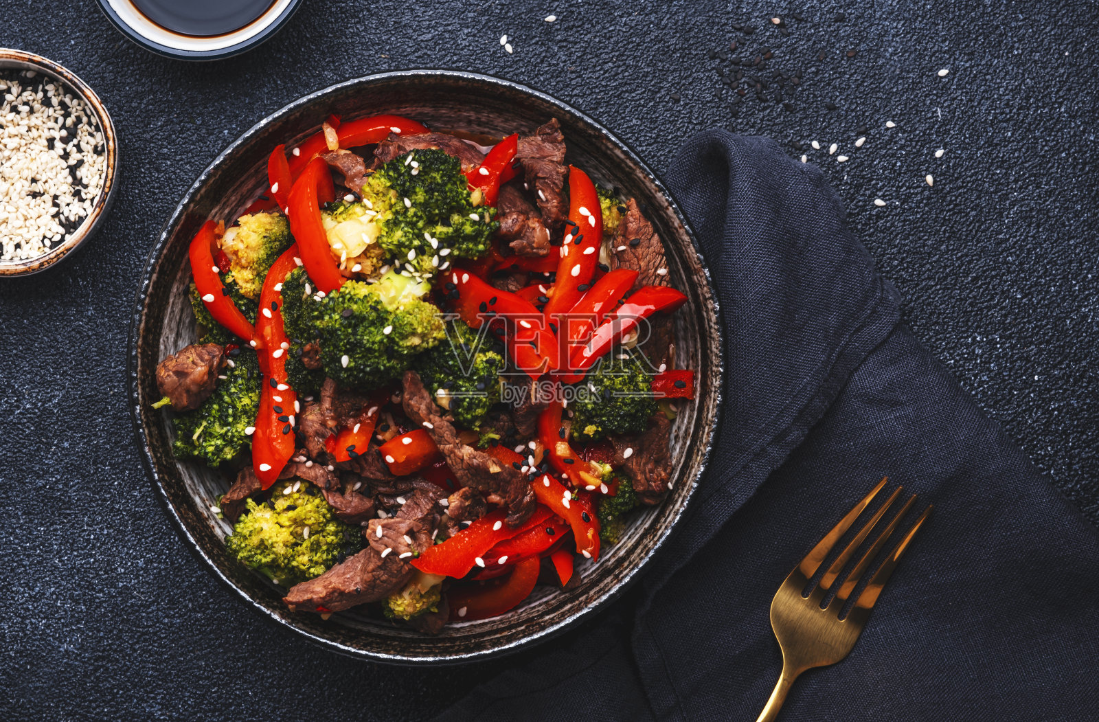 在黑色的桌子背景上，用陶瓷碗翻炒牛肉片、红辣椒和芝麻花椰菜照片摄影图片