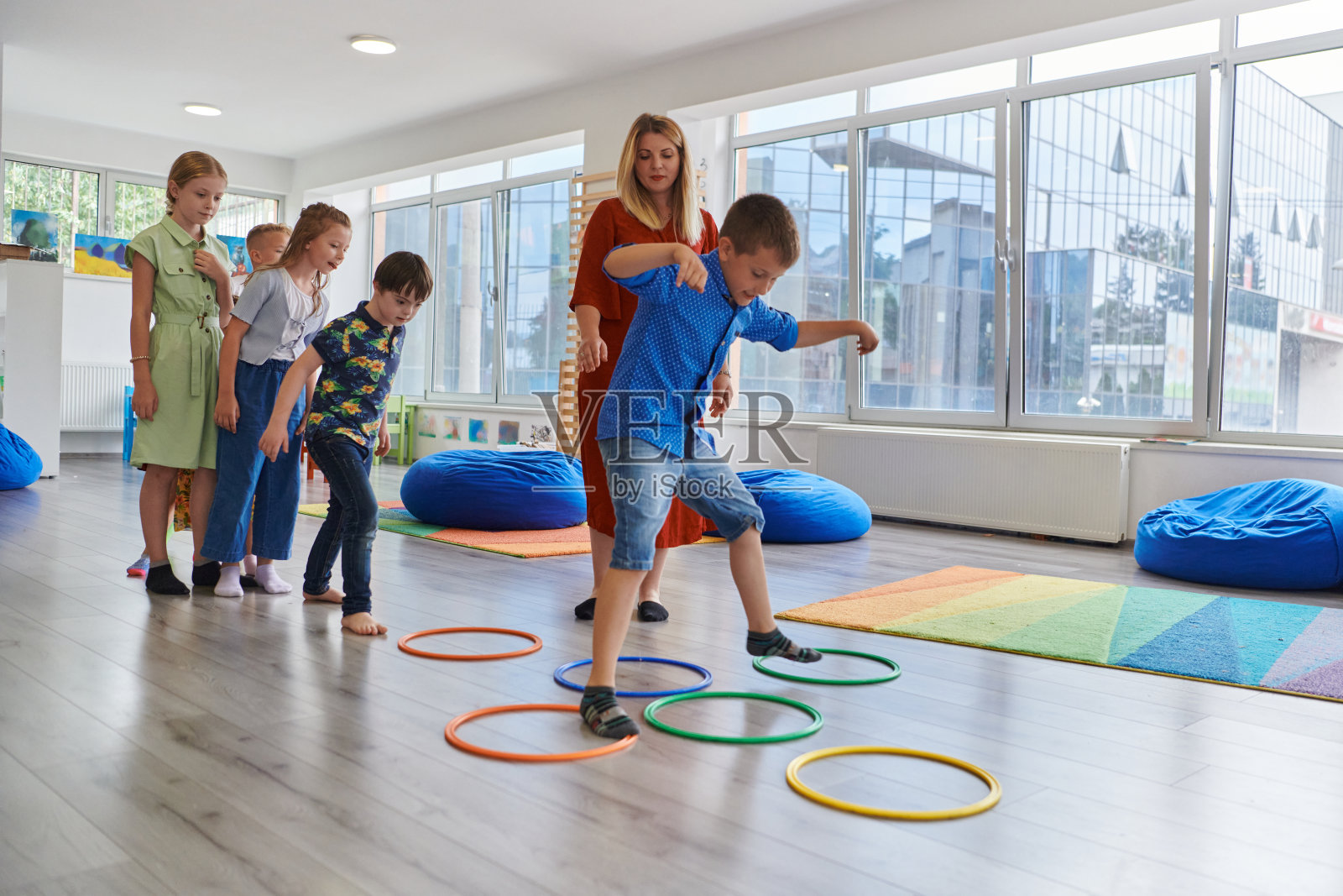 小幼儿园的孩子们带着女老师在教室室内地板上做运动。跳过呼啦圈在地板上绕圈。照片摄影图片