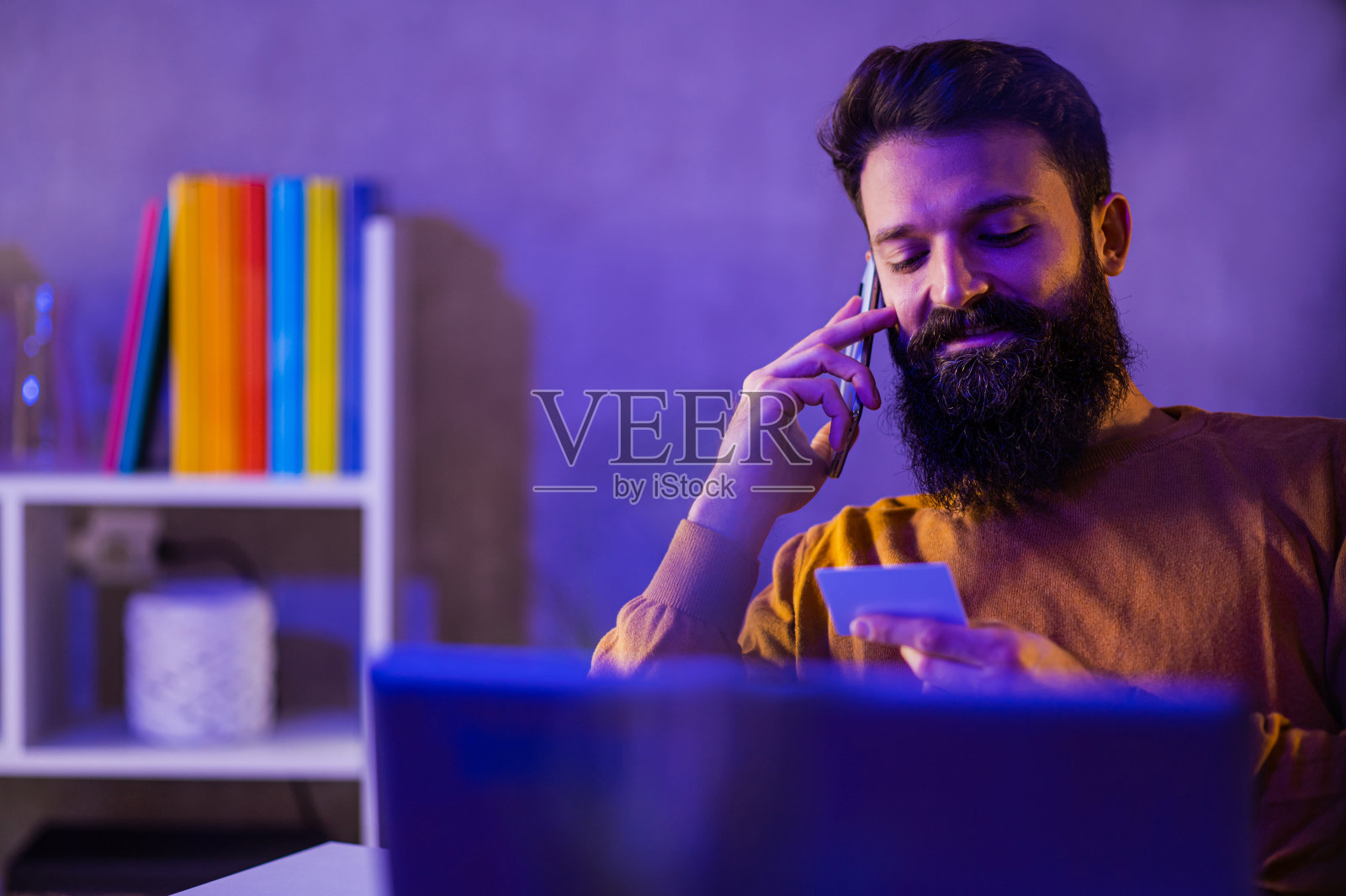 一个快乐的潮人坐在蓝色和紫色霓虹灯的家庭办公室里，拿着信用卡打电话给银行。照片摄影图片