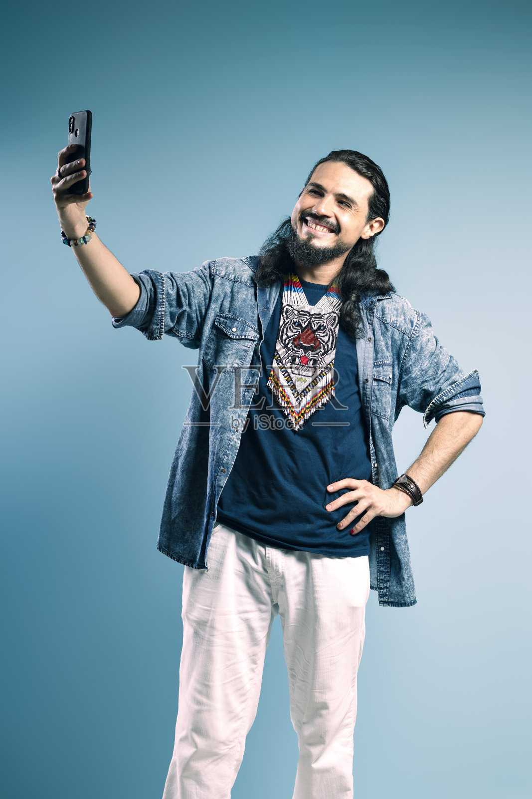 一个巴西人的肖像，穿着纽扣衬衫和牛仔裤，一只手放在腰上，另一只手拿着手机，微笑着自拍——bel<s:1> - par<e:1> -巴西照片摄影图片