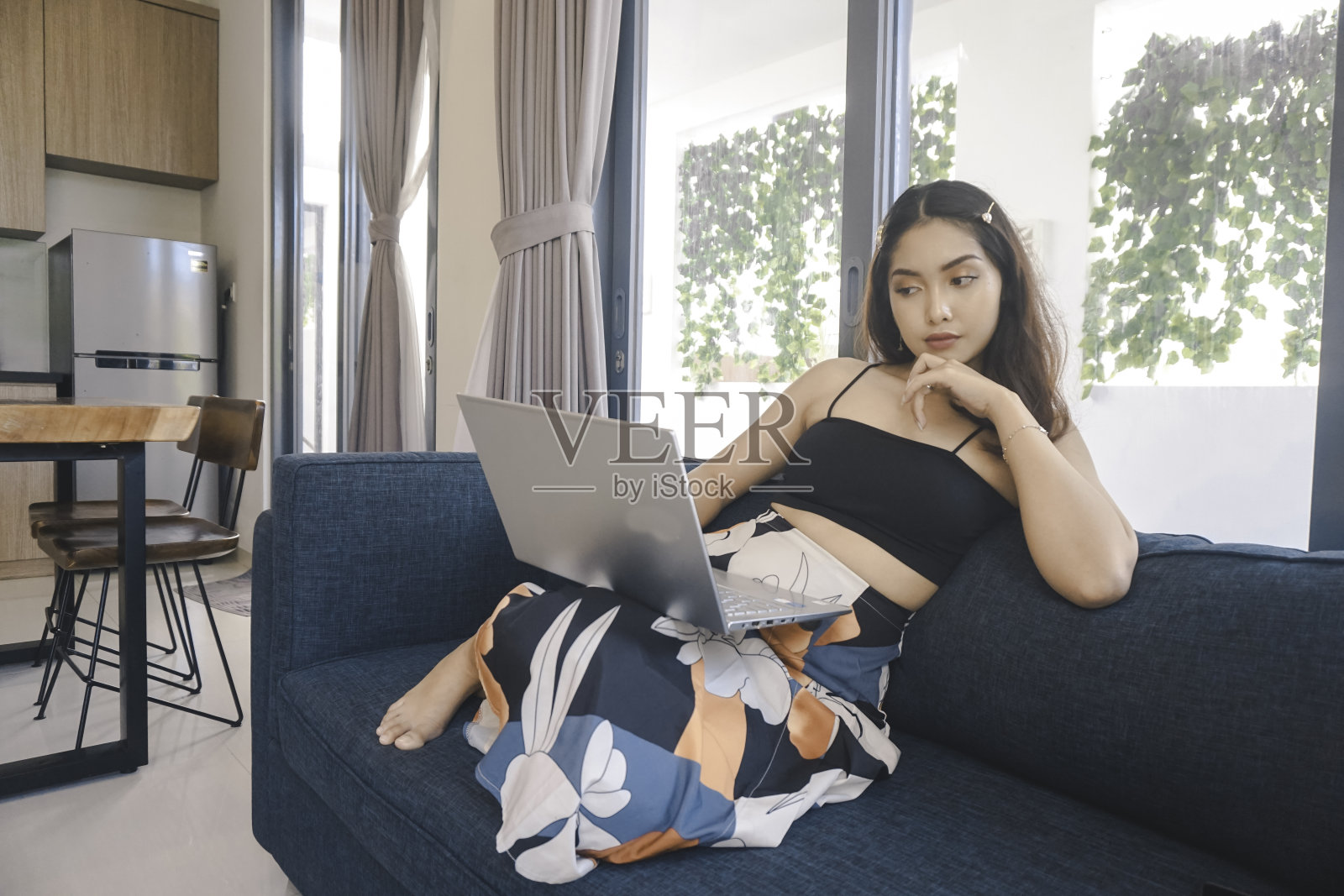 年轻的亚洲女性在家里坐在客厅舒适的沙发上思考和用笔记本电脑工作。照片摄影图片