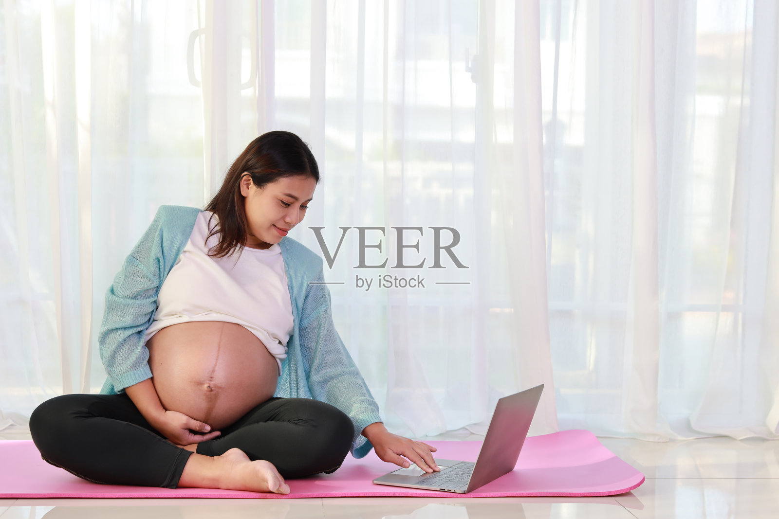 快乐的年轻亚洲孕妇坐在瑜伽垫上练习冥想瑜伽，在家里客厅的电脑上观看在线训练视频。准妈妈在怀孕期间为孩子的出生做准备照片摄影图片