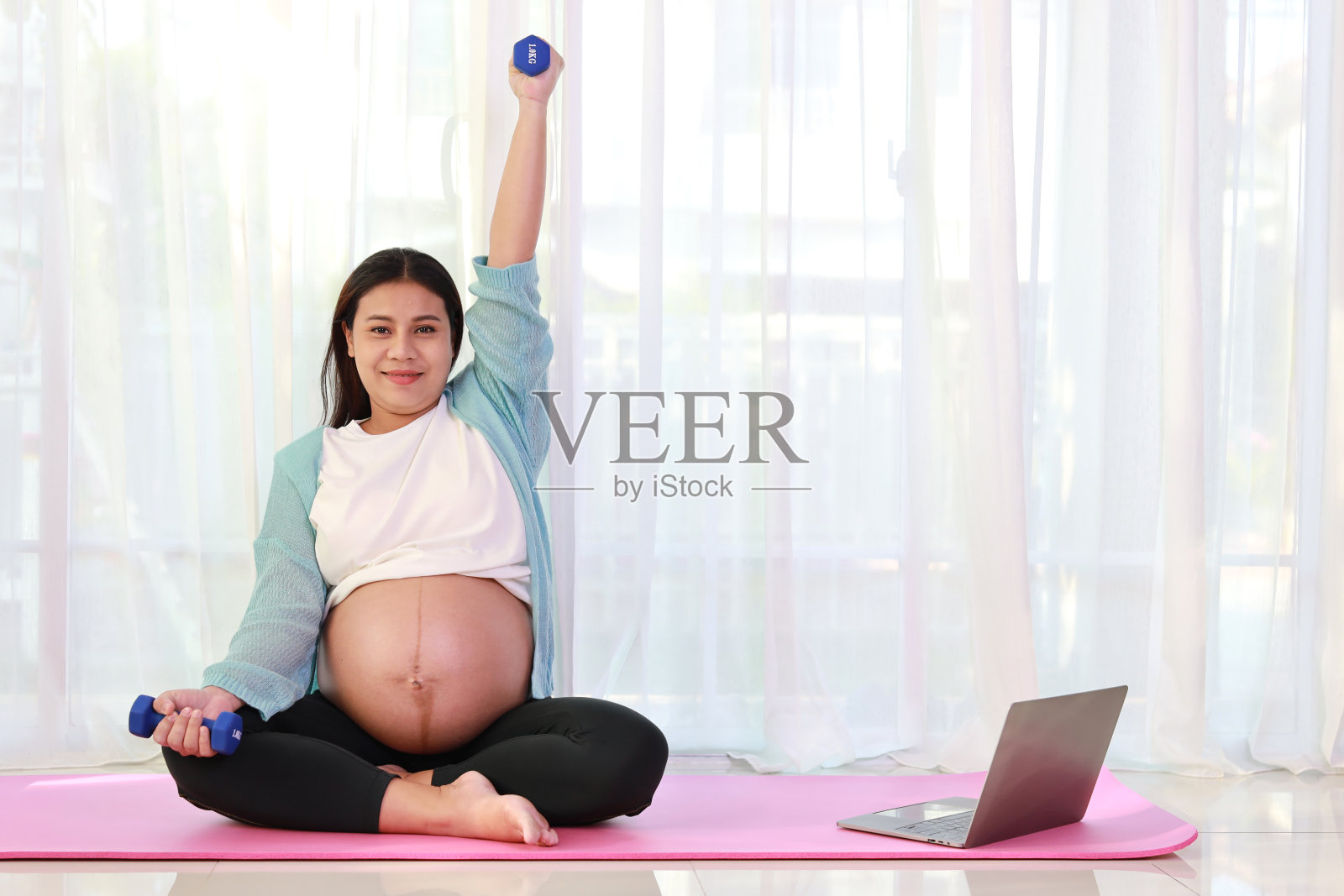 快乐的年轻亚洲孕妇坐在粉红色的垫子上举哑铃，在家里客厅的电脑上观看在线培训视频。准妈妈在怀孕期间为孩子的出生做准备照片摄影图片