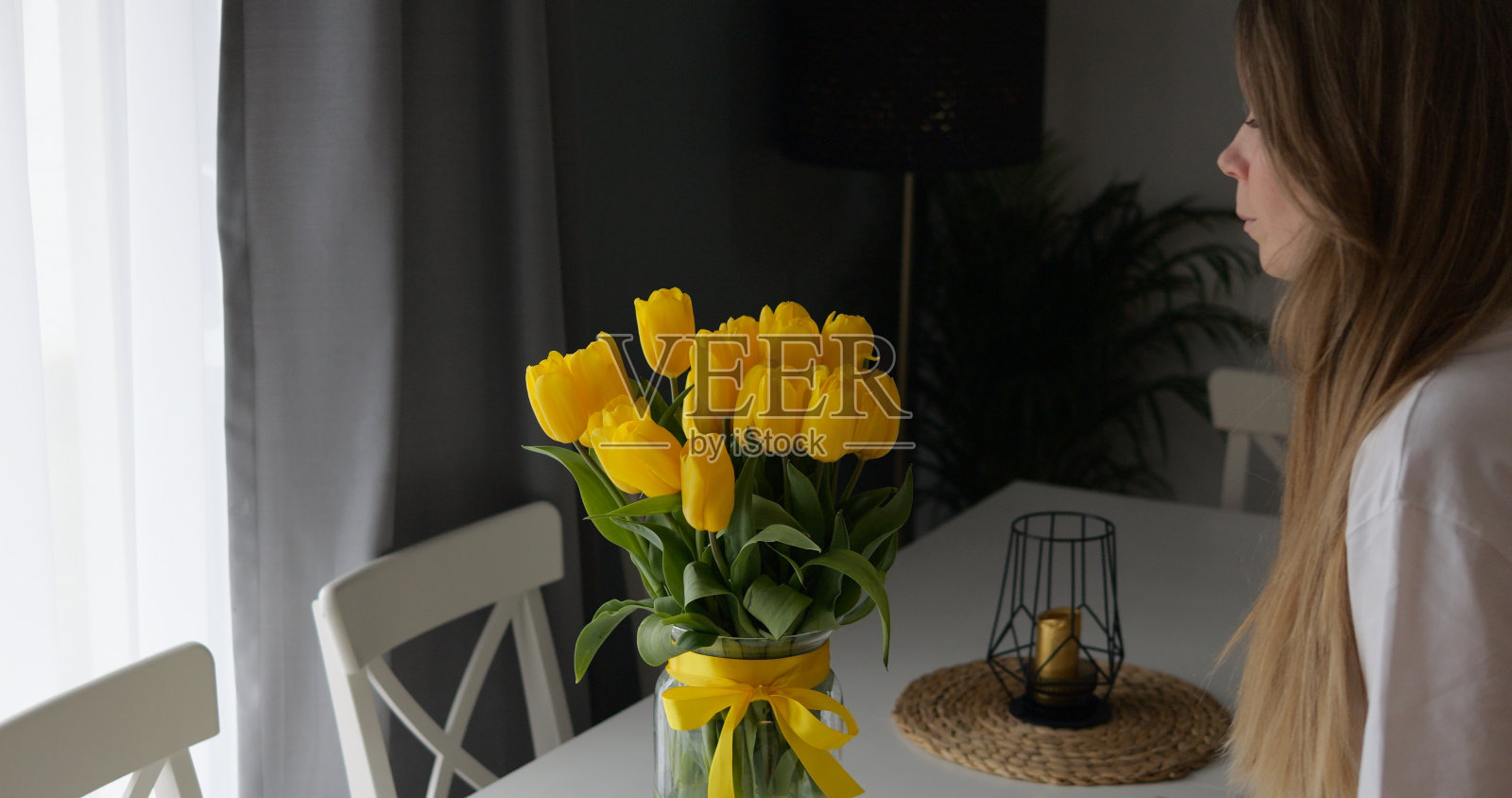 年轻漂亮的女士看着桌子上一束明亮的黄色郁金香。家中散发着浪漫的气息和气氛，玻璃花瓶上装饰着蝴蝶结和蝴蝶结。照片摄影图片