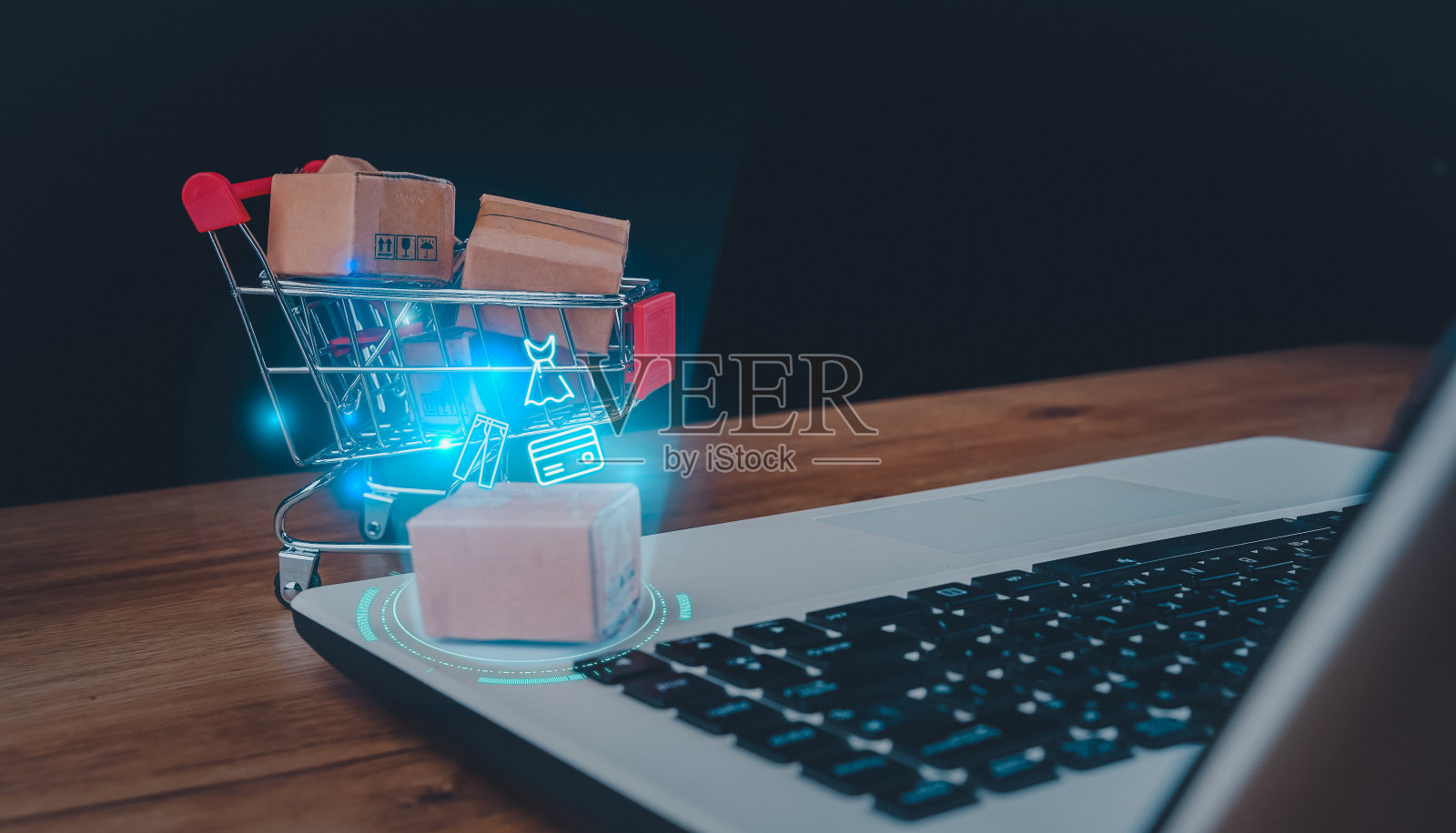 网上购物和送货服务理念。购物车里的棕色纸盒，办公室背景的木桌上放着笔记本电脑键盘。顾客通过互联网从零售商网站订购商品。照片摄影图片