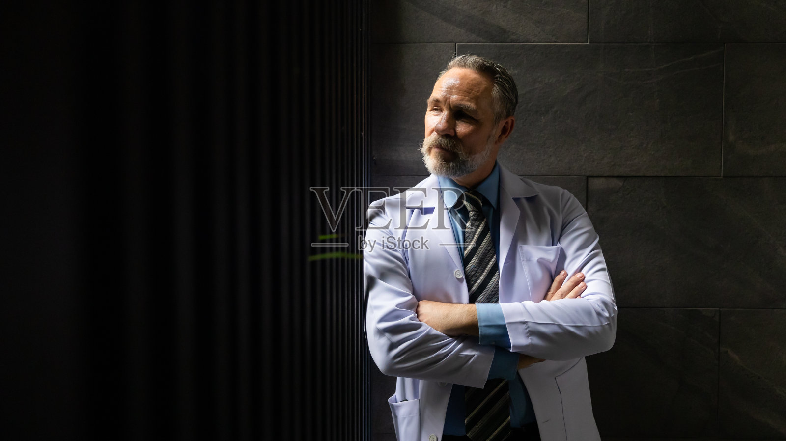 强烈的反差和戏剧性的概念。肖像白人老年男子医生摆姿势和手臂交叉在健康中心和医院。医疗保健和医药照片摄影图片