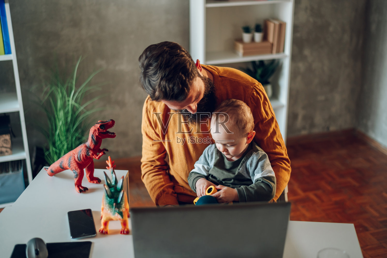 在现代家庭办公室里，一个孩子坐在父亲的腿上玩玩具。照片摄影图片