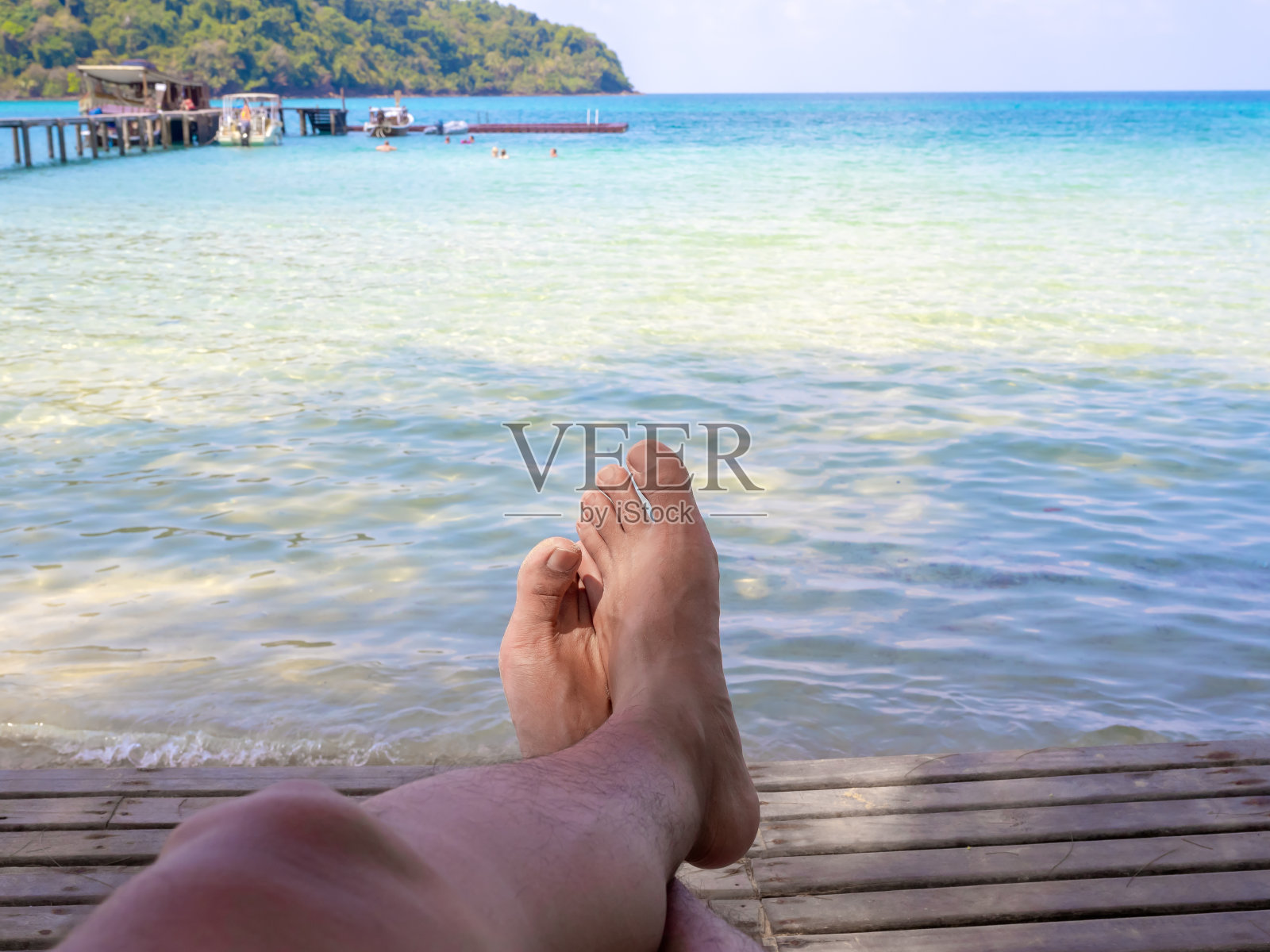 沙滩上男人的腿。男躺在沙滩上的木椅上，以蔚蓝的天空和大海为背景，在夏日的阳光灿烂的日子里，在海边放松，假日度假旅行。照片摄影图片