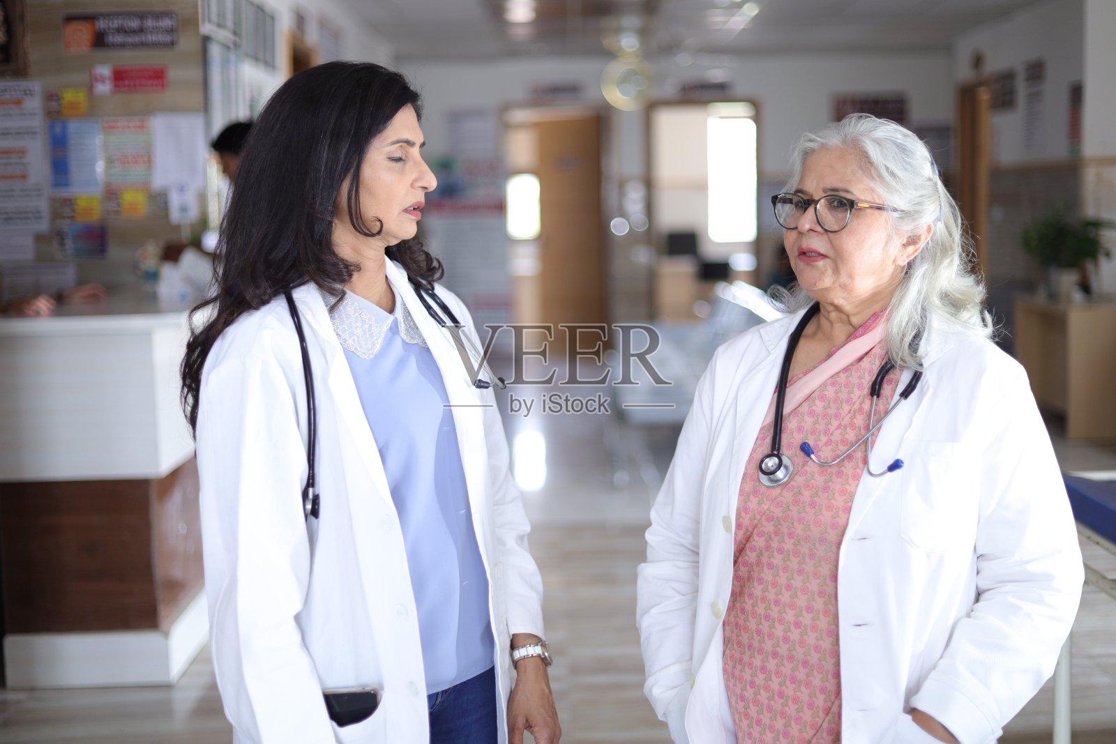 印度医生与助手讨论。医生们在走廊里谈笑风生。年长的女医生和年轻的医生。医生在医院阅读病人的书面报告或医疗文件。照片摄影图片