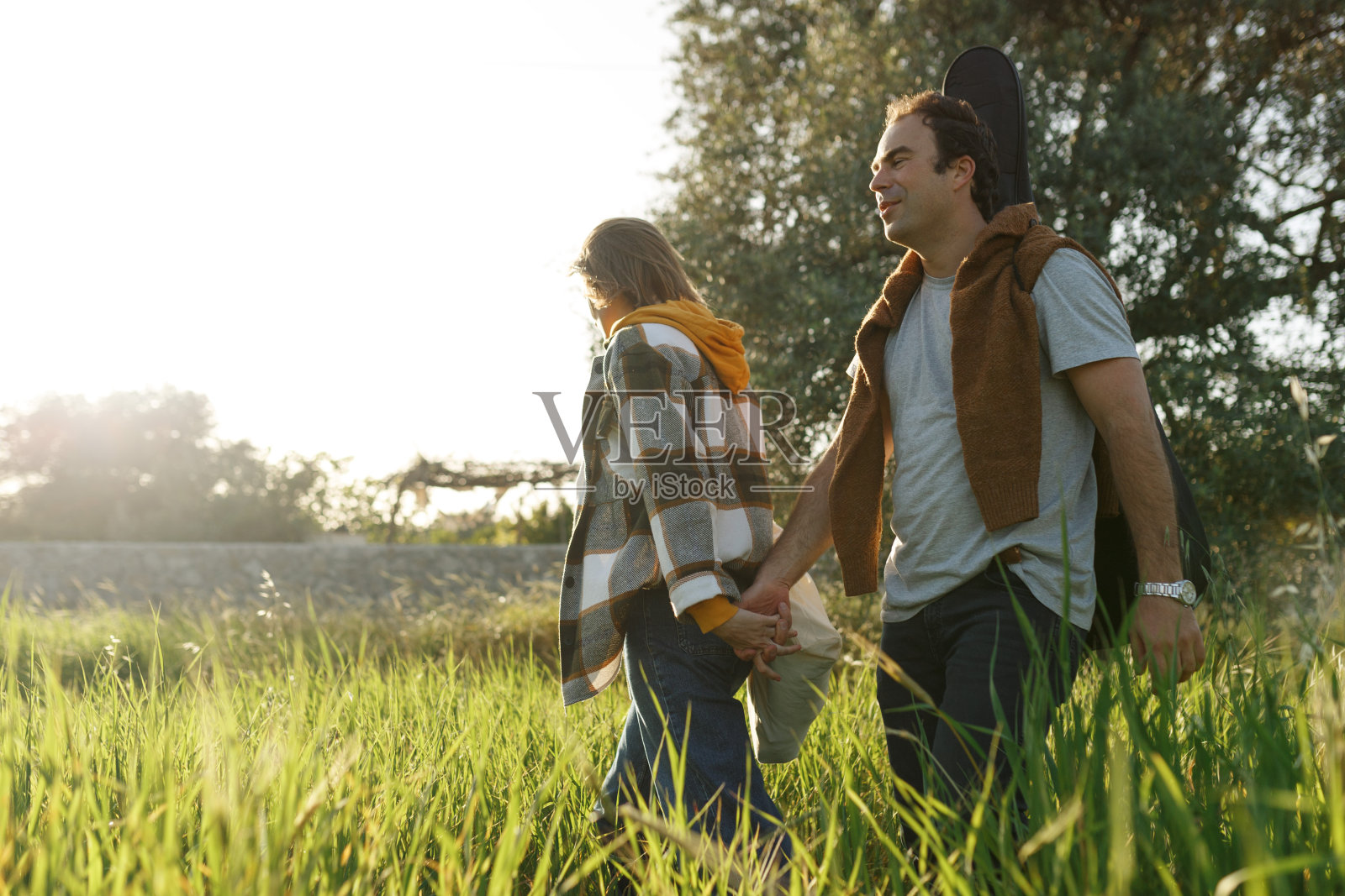 一对年轻的中年夫妇在公园里愉快地散步聊天。夏日公园里的一对成熟夫妇。一男一女，穿着休闲装，拿着吉他和野餐包，在春天的大自然中外出。照片摄影图片
