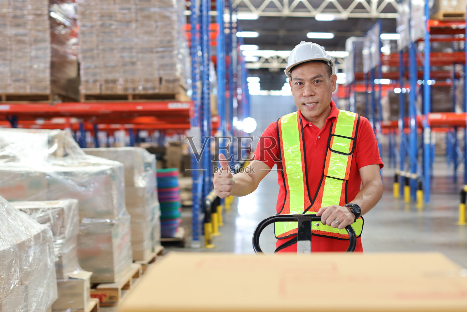 在零售仓库物流配送中心，仓库工人戴着安全帽，穿着反光夹克，拉着托盘卡车，拿着或上传大箱子包裹到货架上照片摄影图片
