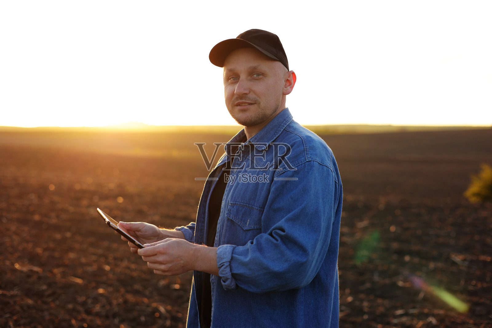 夕阳下，一位微笑着的男性农民在犁地的田地里拿着数码平板电脑，看着相机。检查和控制土壤质量，土地准备播种作物。智能农业技术。照片摄影图片