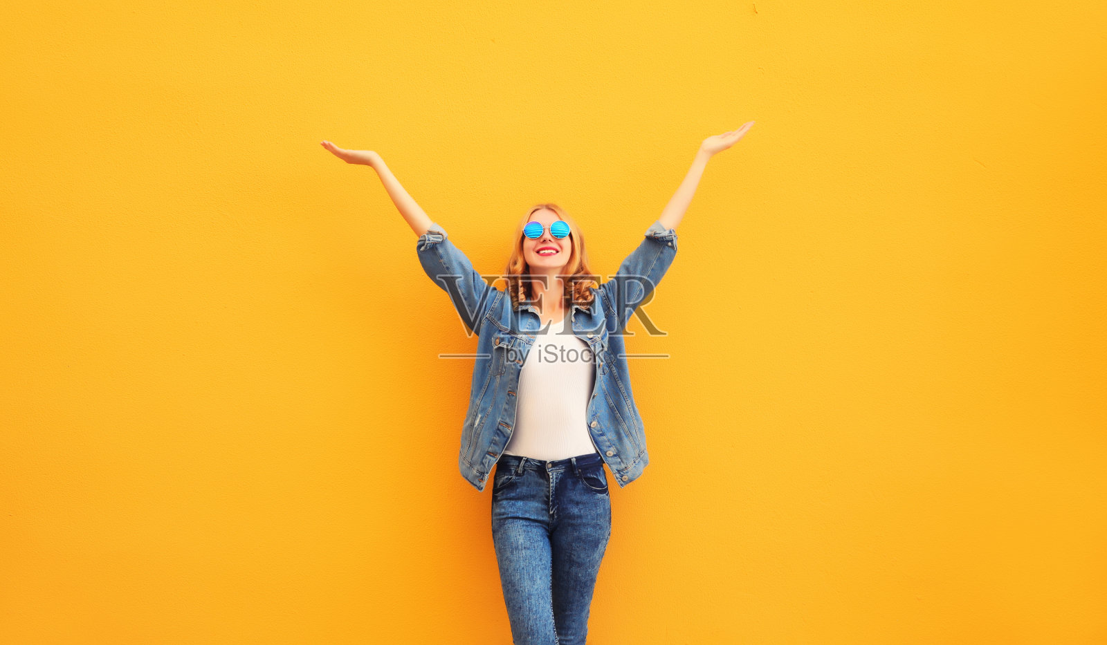 快乐开朗的白人微笑的年轻女子举起她的手，穿着黄色背景的牛仔夹克照片摄影图片