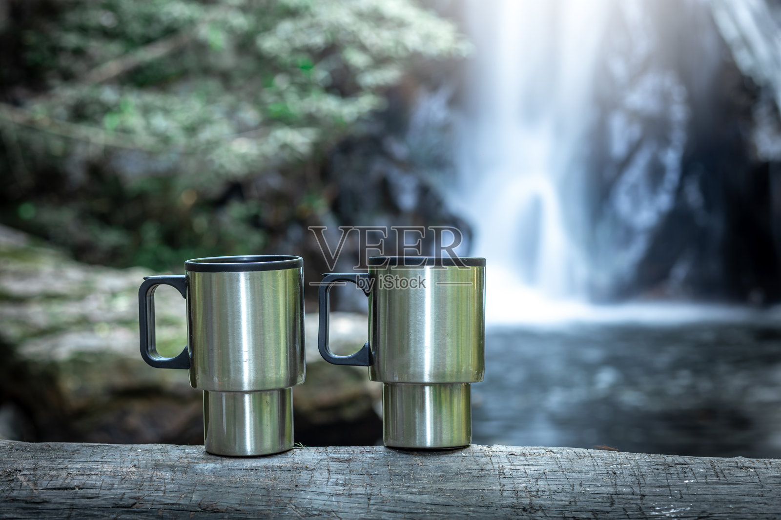 靠近两个铁咖啡杯在木与瀑布背后。照片摄影图片