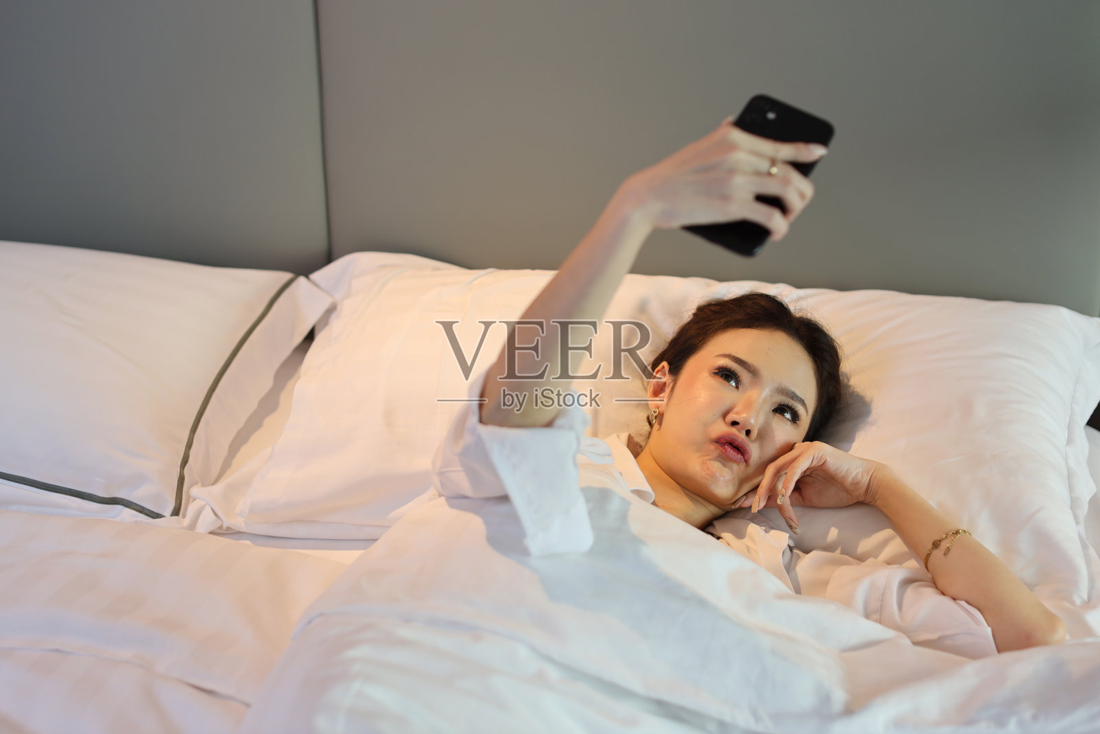 俯视图年轻美丽的亚洲女人穿着白色睡衣躺在床上自拍，在做好梦之前用手机打招呼。生活方式的概念照片摄影图片