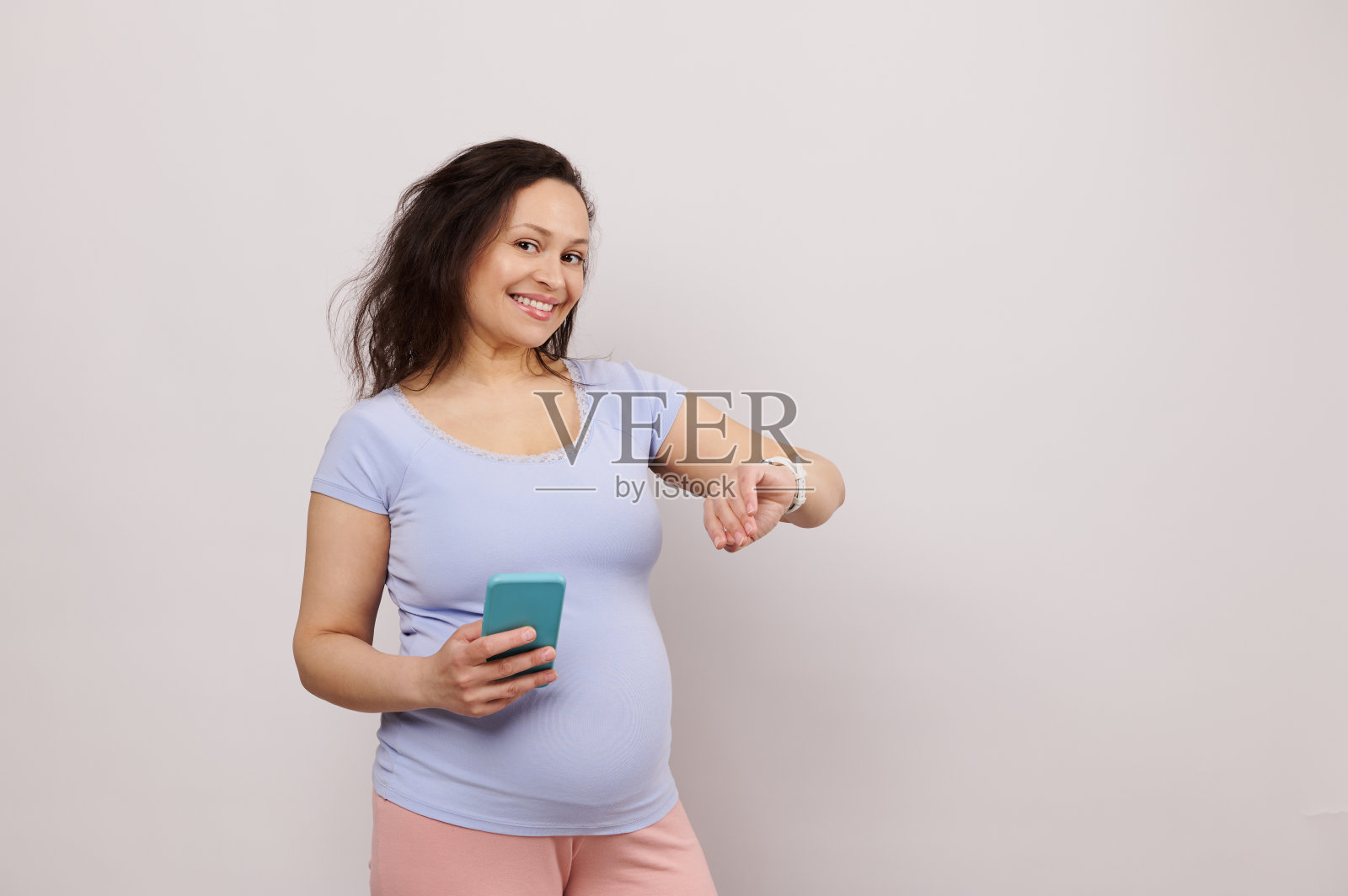 快乐微笑的孕妇期待着孩子，拿着智能手机，检查电子智能手表上的移动应用程序。照片摄影图片