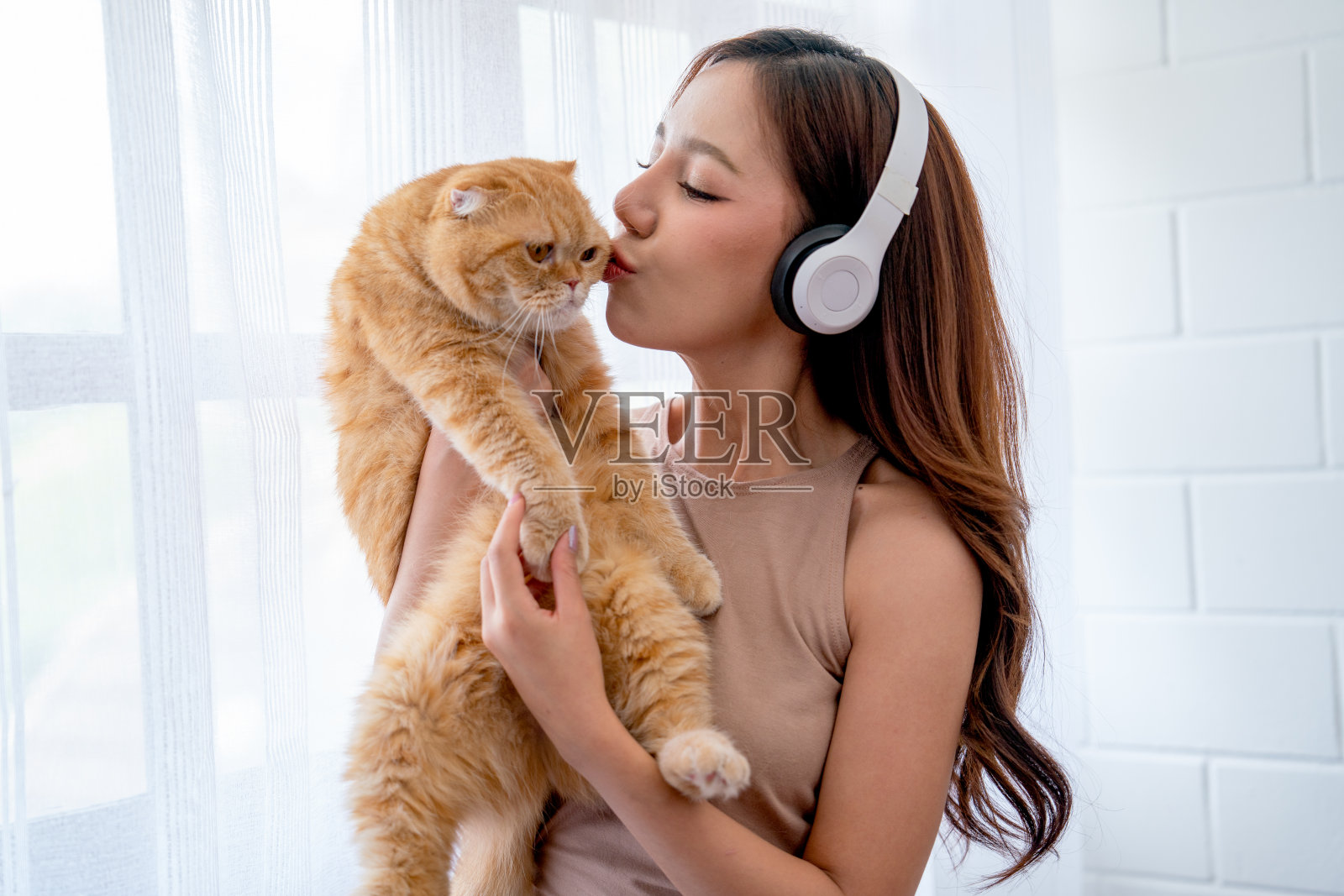 漂亮的亚洲女孩戴着耳机，抱着猫在白色窗帘的玻璃窗前亲吻，他们在家里看起来很幸福。照片摄影图片